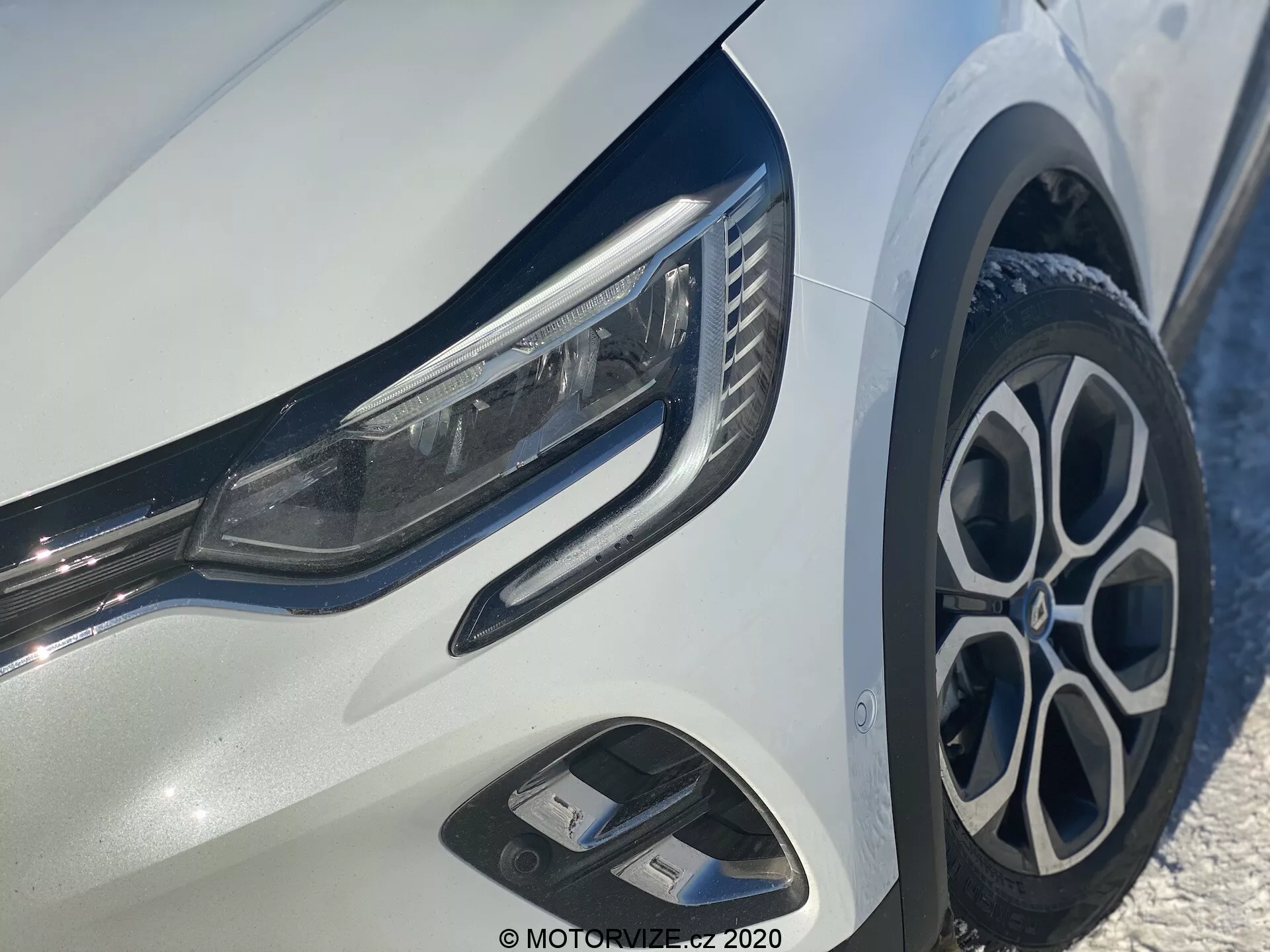 Zbliżenie na Renault Captur 2020, skupiające się na przedniej lewej ćwiartce, pokazujące eleganckie reflektory LED, stylowe aluminiowe felgi wieloramienne, dynamiczny przedni zderzak z wlotami powietrza i błyszczące chromowane akcenty na jasnym białym nadwoziu.