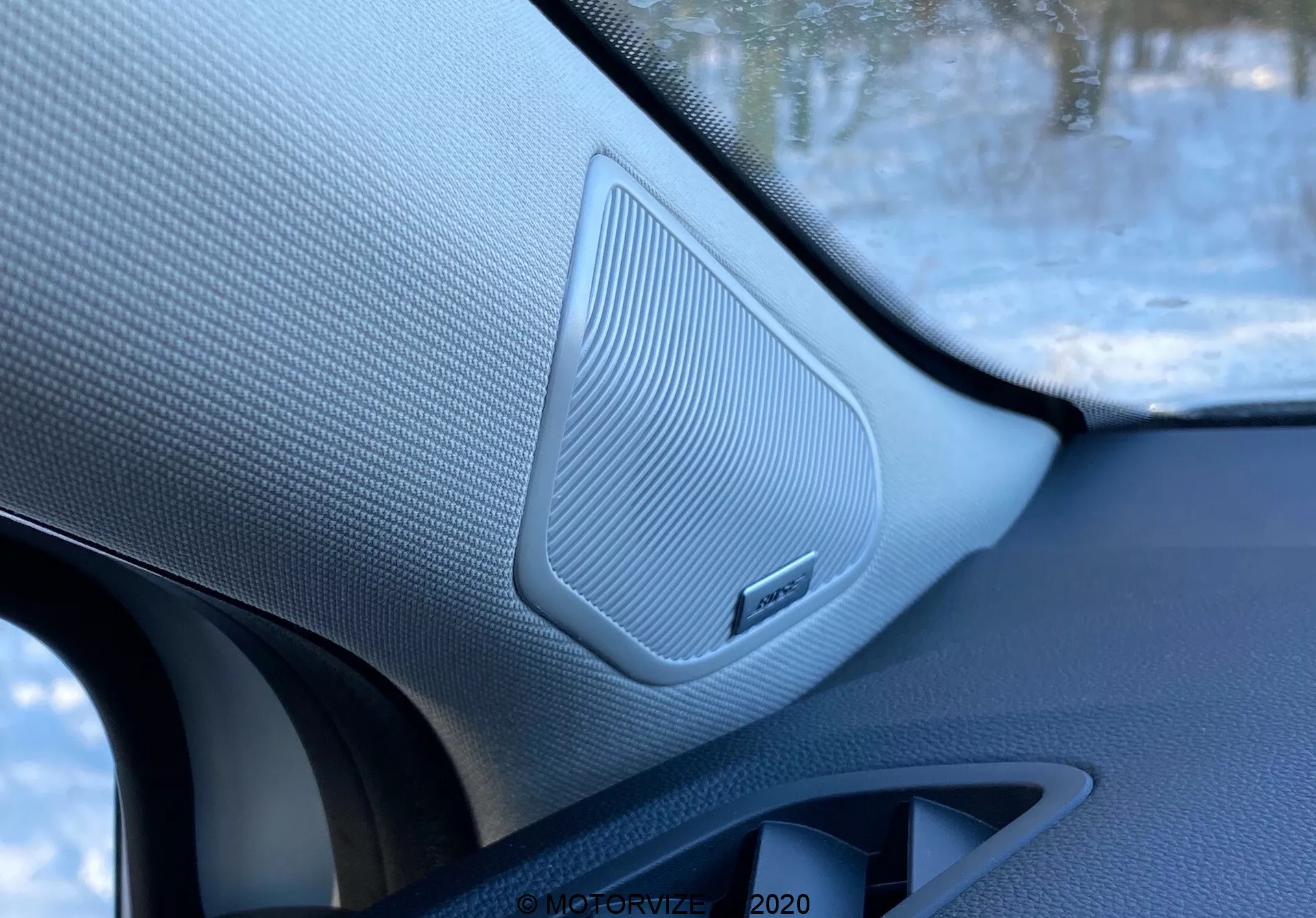 Zbliżenie wnętrza Renault Captur 2020, przedstawiające głośnik na przednich drzwiach pasażera z teksturowaną, siateczkową maskownicą, otoczony kontrastującymi materiałami i emblematem marki, zrobione z wnętrza pojazdu.