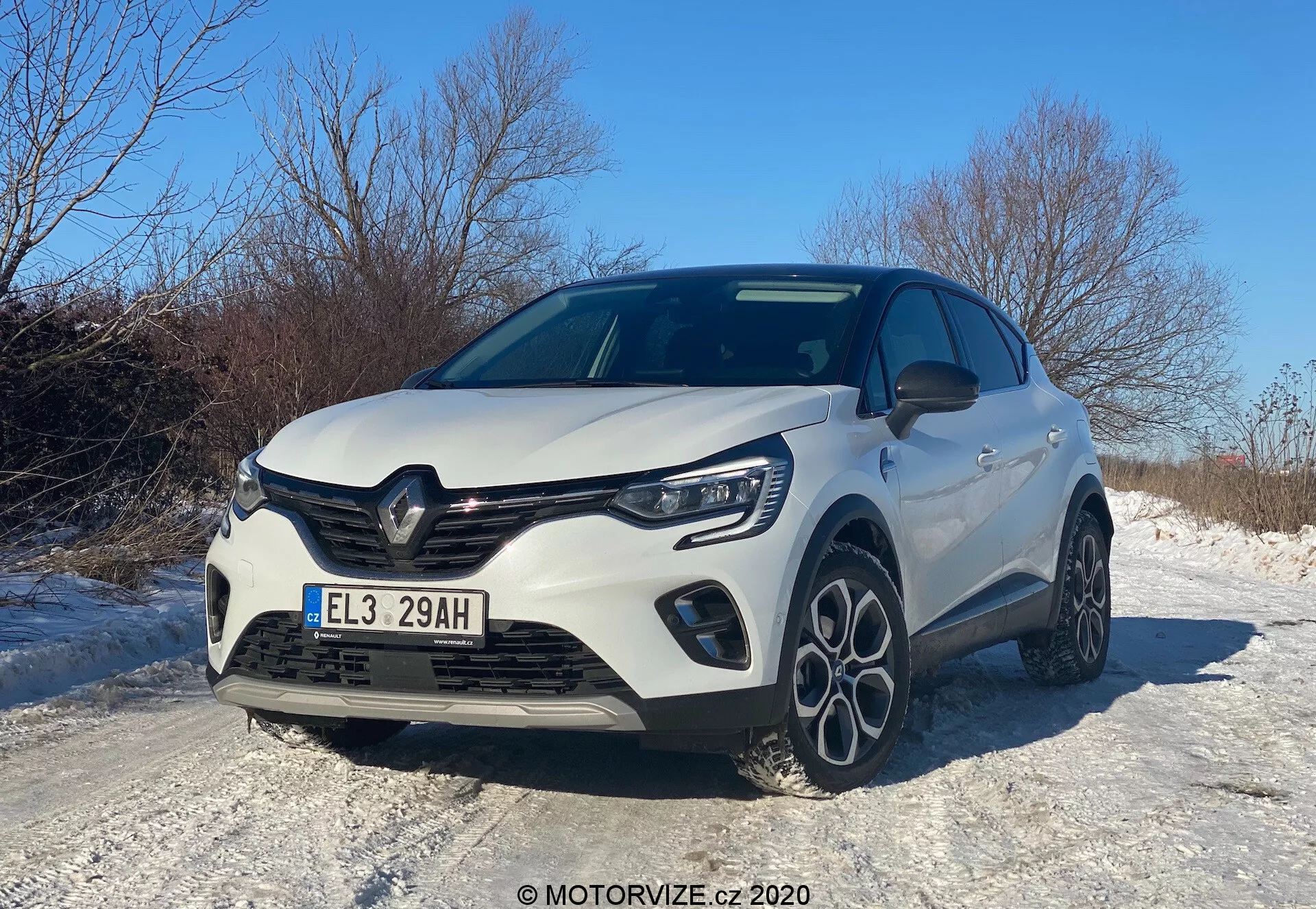 Nowoczesny hatchback Renault Captur 2020 uchwycony z niskiego przedniego trójkątnego kąta, prezentujący przednią kratkę, maskę, reflektory LED, przedni zderzak z zintegrowanymi światłami przeciwmgielnymi oraz częściowy profil boczny. Elementy designu obejmują wyraźną kratkę z emblematem producenta, rzeźbione linie na masce i wieloramienne felgi aluminiowe, wszystko to pod jasnym dziennym niebem, odzwierciedlając sportową i aerodynamiczną estetykę, prawdopodobnie podkreślając ulepszony poziom wykończenia lub pakiet sportowy. Samochód znajduje się na zaśnieżonej drodze, co sugeruje cechy odpowiednie do jazdy zimowej.