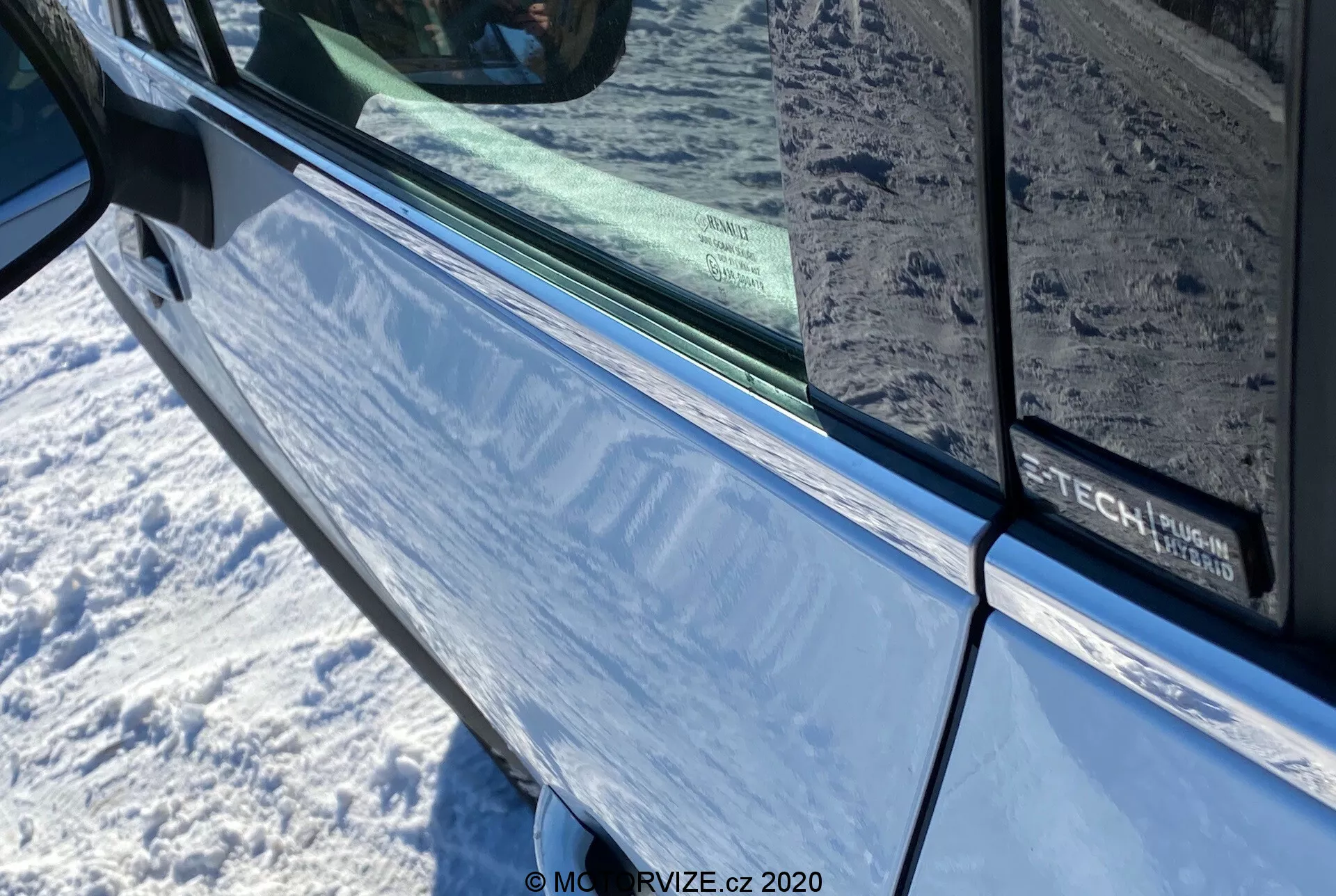 Zbliżenie na Renault Captur 2020 w metalicznym niebieskim, skoncentrowane na drzwiach kierowcy i bocznym lusterku z wbudowanym kierunkowskazem. Zdjęcie podkreśla obszar drzwi z czarną uszczelką biegnącą wzdłuż krawędzi przyciemnianej szyby, odbijającej otoczenie. Na drzwiach widać częściowo napis wskazujący na możliwy pakiet wyposażeniowy lub technologiczny.