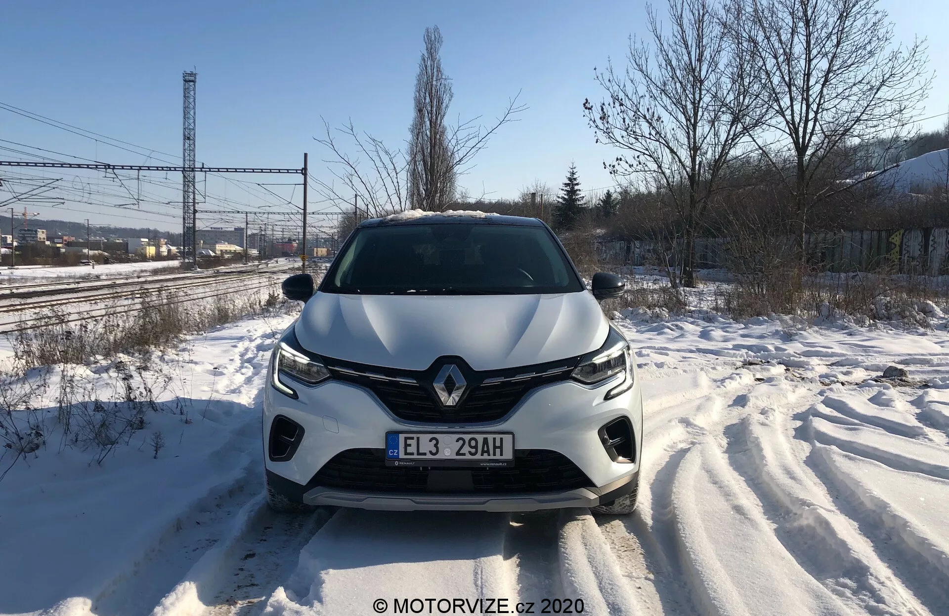 Bezpośredni widok z przodu na Renault Captur 2020 na zaśnieżonej drodze, ukazujący szczegółowo przedni grill, reflektory, rzeźbioną maskę i przedni zderzak w ciągu dnia. Widoczne ślady opon świadczą o niedawnym ruchu. Design pojazdu sugeruje aerodynamiczne kształty, z wyraźnym emblematem marki i zaawansowanym systemem oświetlenia. W przednim zderzaku mogą być zintegrowane czujniki lub kamery asysty kierowcy.