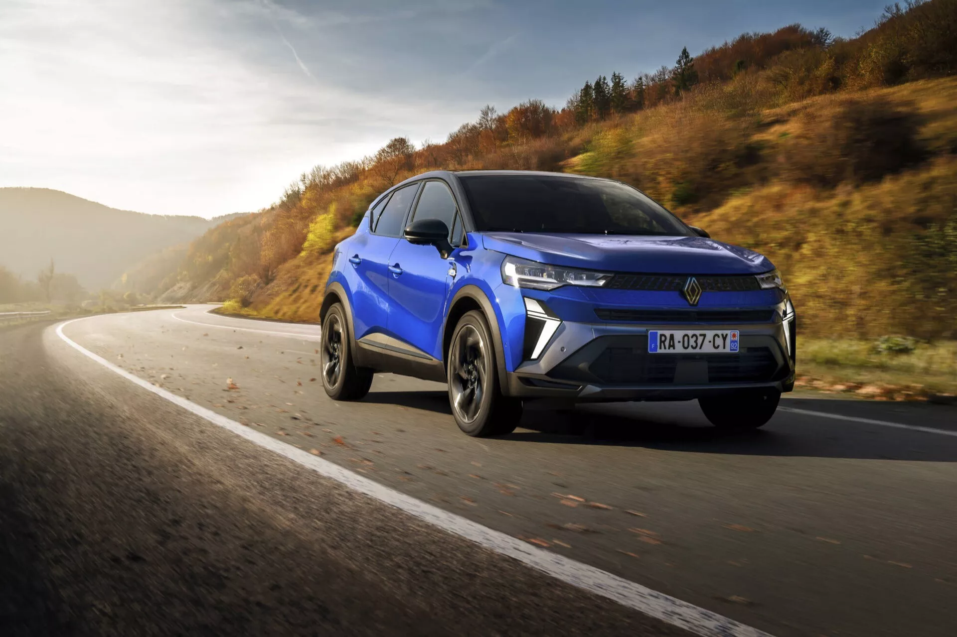 Niebieskie SUV Renault Captur (II Facelift) 2024 w dynamicznym ruchu na zakrzywionej asfaltowej drodze, prezentujące fasadę przednią i profil boczny pod ciepłym światłem słonecznym