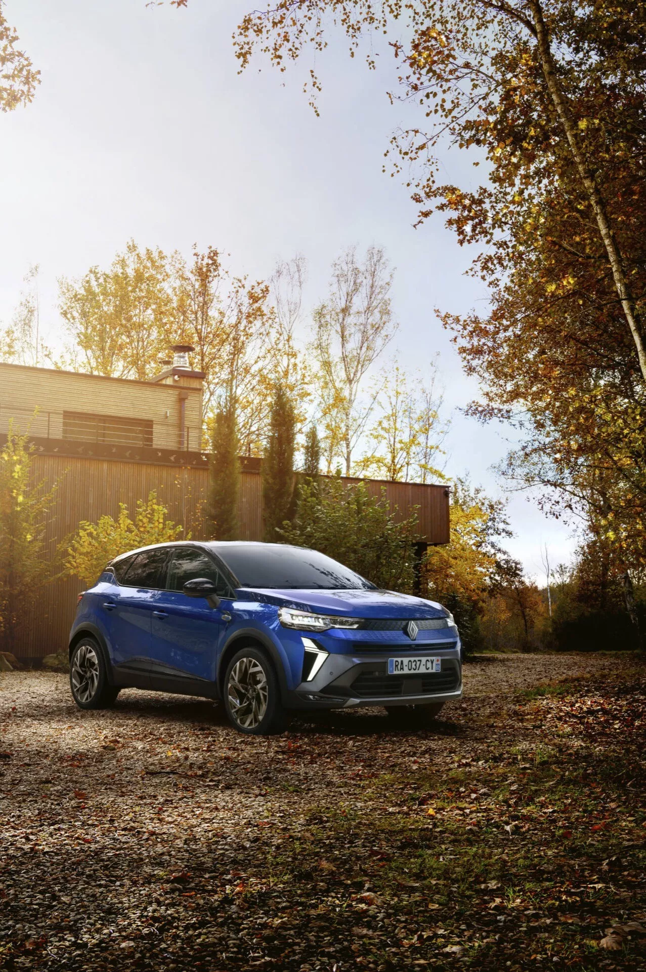 Model Renault Captur II lifting 2024, niebieskie kompaktowe SUV zaparkowane na zewnątrz otoczone jesiennym liśćmi, pokazujące dominujący i kątowy widok z lewej przedniej strony z detalami projektu, takimi jak ostre linie, charakterystyczne reflektory i felgi aluminiowe, podkreślające potencjalne cechy jak panoramiczne okno dachowe i nowoczesna technologia w jesiennym otoczeniu