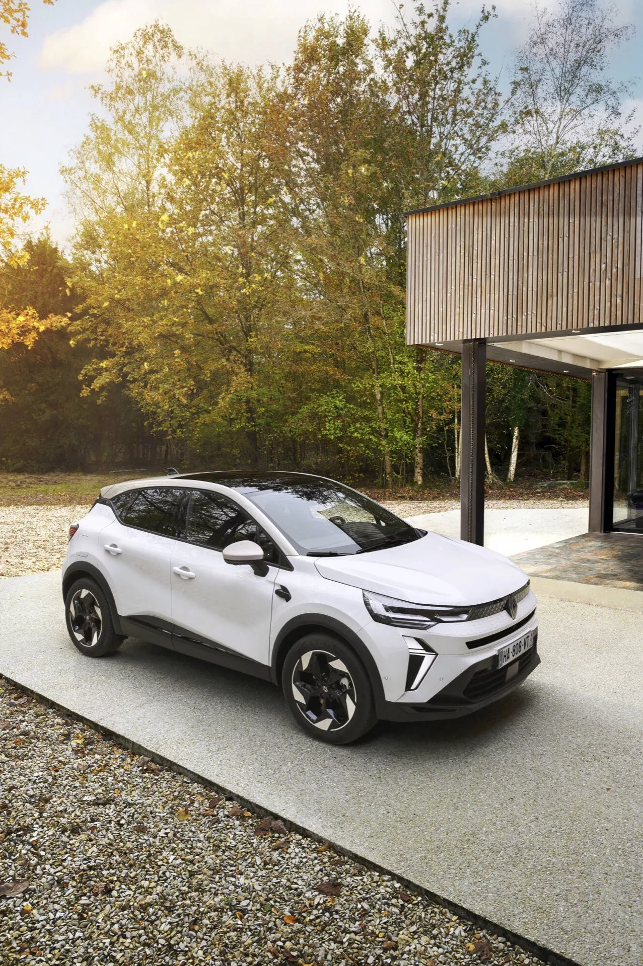 Facelifting Renault Captur 2024 uchwycony z wysokiego kąta na zewnątrz, możliwie w leśnym otoczeniu, podkreślający nowoczesny design pojazdu i cechy technologiczne, takie jak reflektory LED, dwukolorowa paleta barw i aerodynamiczne koła, sugerujący ekologiczny model elektryczny lub hybrydowy.