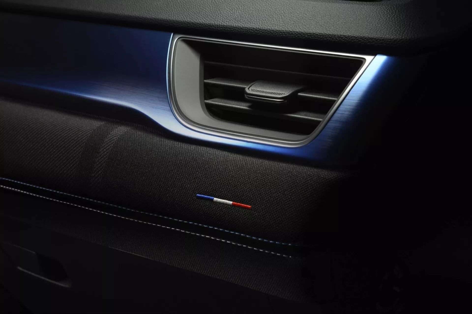 Szczegół wnętrza Renault Captur II facelift 2024 przedstawiający panel drzwiowy z elegancką metalową klamką, delikatne niebieskie oświetlenie ambientowe, premium wkładkę drzwiową ze szwami i emblematem flagi narodowej wskazującym na specjalną edycję lub pakiet sportowy