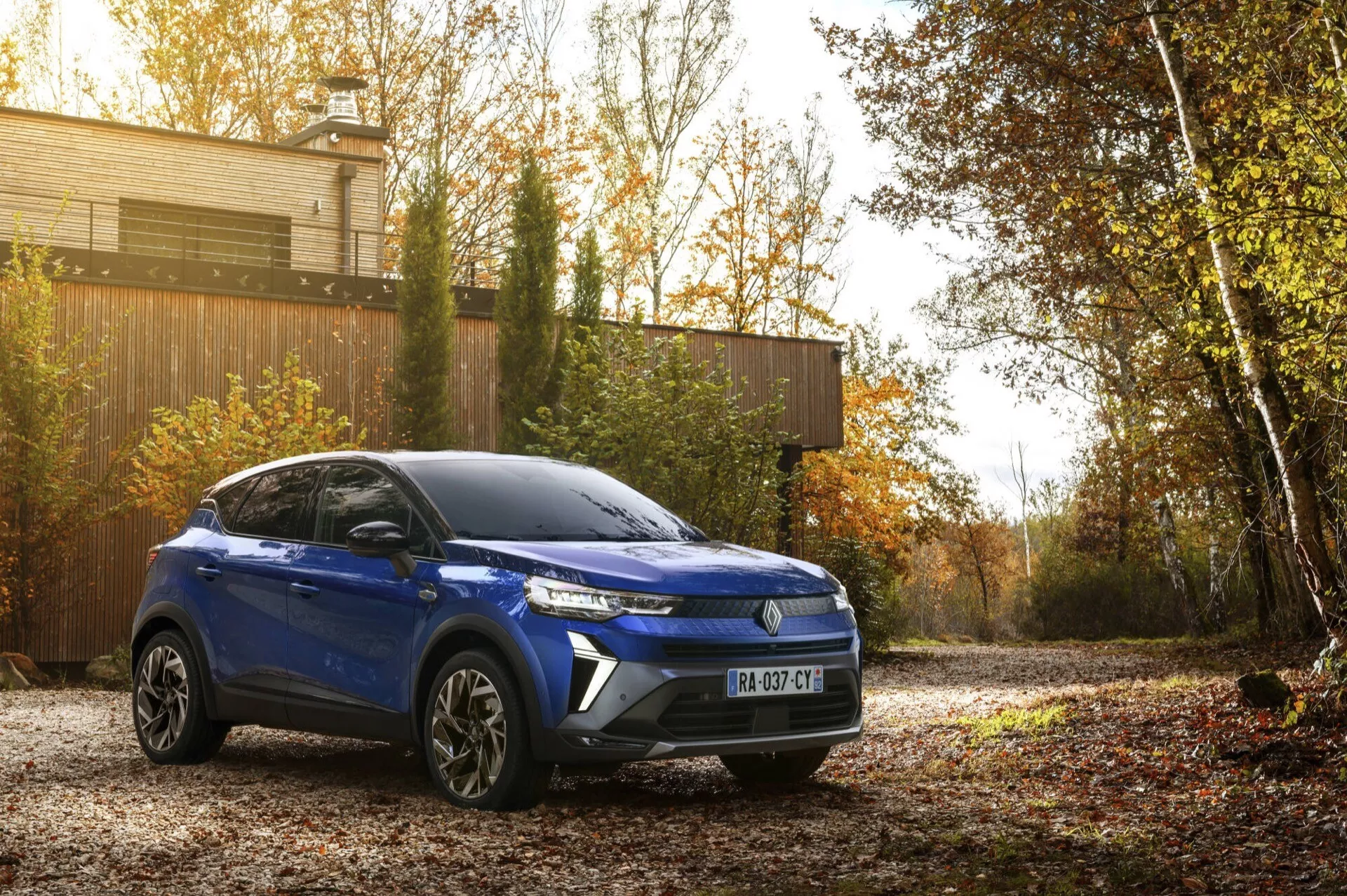 Niebieski Renault Captur II lifting 2024, kompaktowy SUV zaparkowany na zewnątrz prezentujący swój prawo-przedni kwartał z naciskiem na nowoczesne linie designu, charakterystyczną przednią kratę, reflektory LED i felgi aluminiowe, na jesiennym tle pod naturalnym światłem porannym lub późnym popołudniowym, co podkreśla jego estetyczny urok i zaawansowanie technologiczne