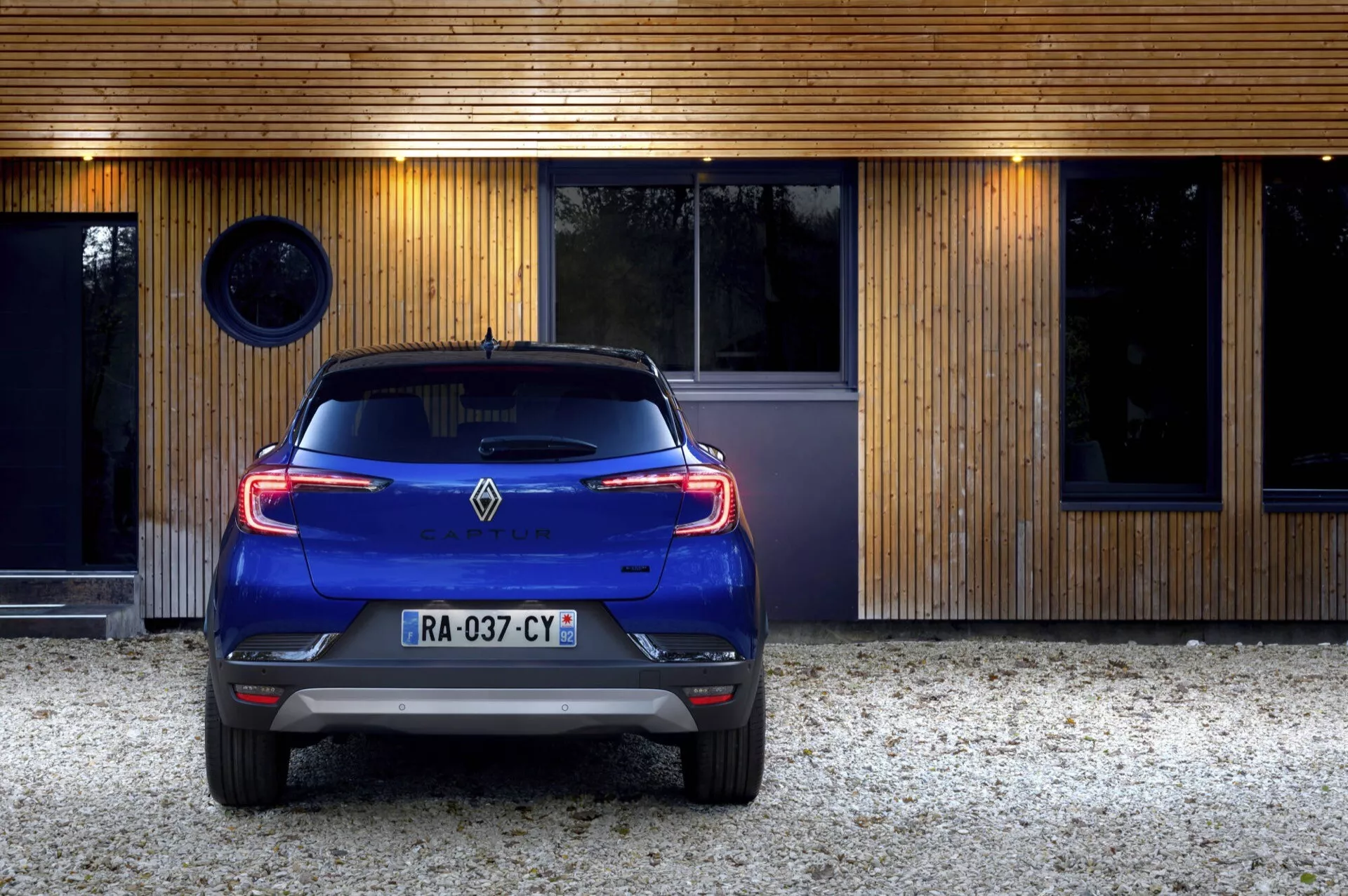 Tylny widok niebieskiego Renault Captur 2024 (II Facelifting) z tylnymi światłami LED zaparkowanego przed nowoczesnym budynkiem mieszkalnym, wyświetlającego czujniki parkowania, podwójny wydech i antenę w kształcie płetwy rekina
