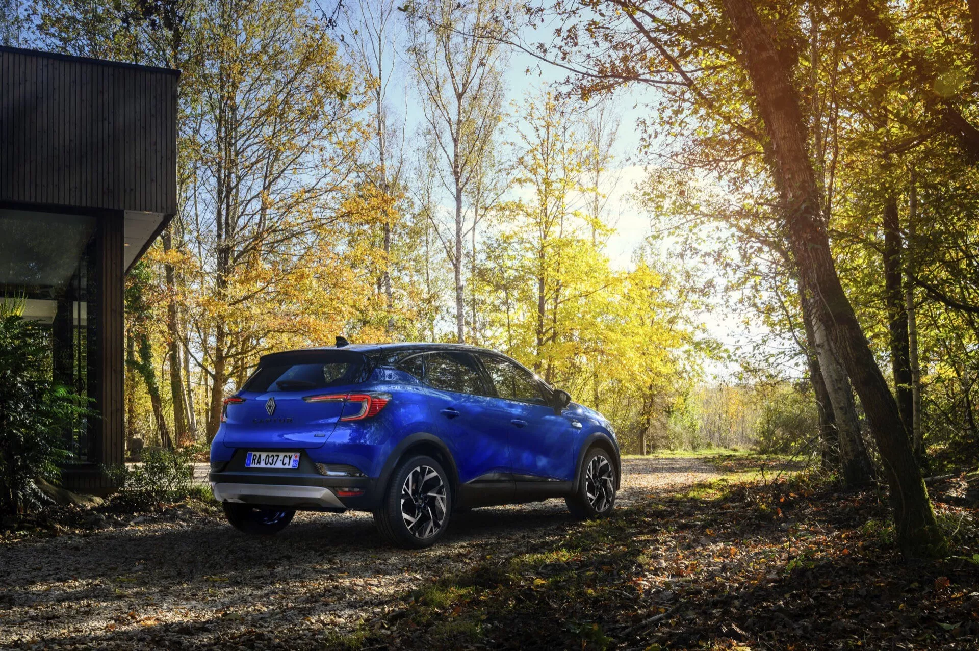 Niebieskie kompaktowe SUV Renault Captur II Facelift 2024 zaparkowane na zewnątrz na nieutwardzonej drodze, prezentujące tylne i boczne profile z tylnymi światłami LED, felgami ze stopu, czujnikami parkowania i zawieszeniem do lekkiej jazdy w terenie w spokojnym lesie jesienią z słońcem przedzierającym się przez drzewa.