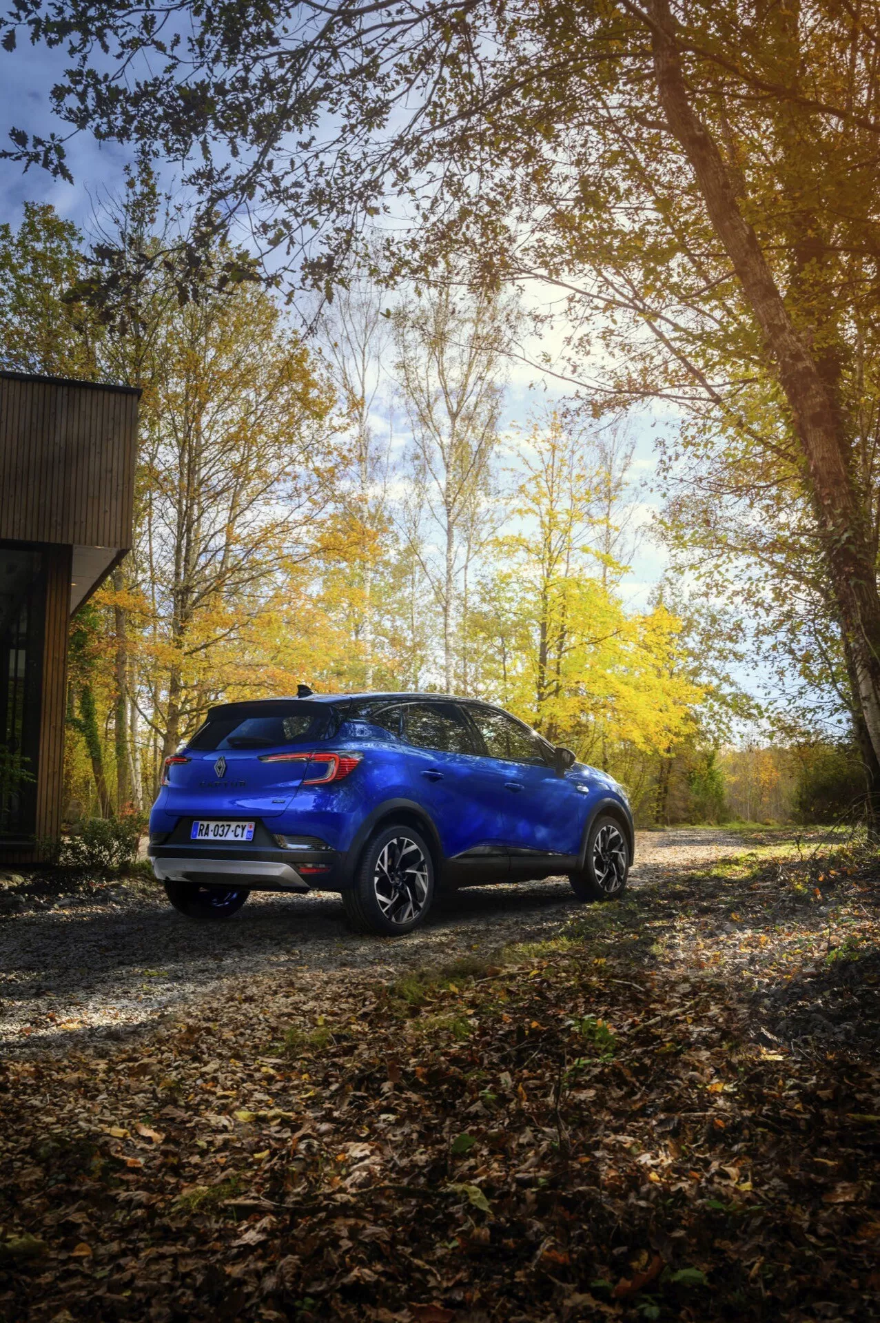 Model Renault Captur II facelifting 2024 w kolorze niebieskim, prezentujący swój tylny i boczny profil na zewnątrz na żwirowej powierzchni z jesienno-leśnym tłem, podkreślający jego linie projektowe, błyszczące wykończenie, felgi aluminiowe i tylny spoiler.