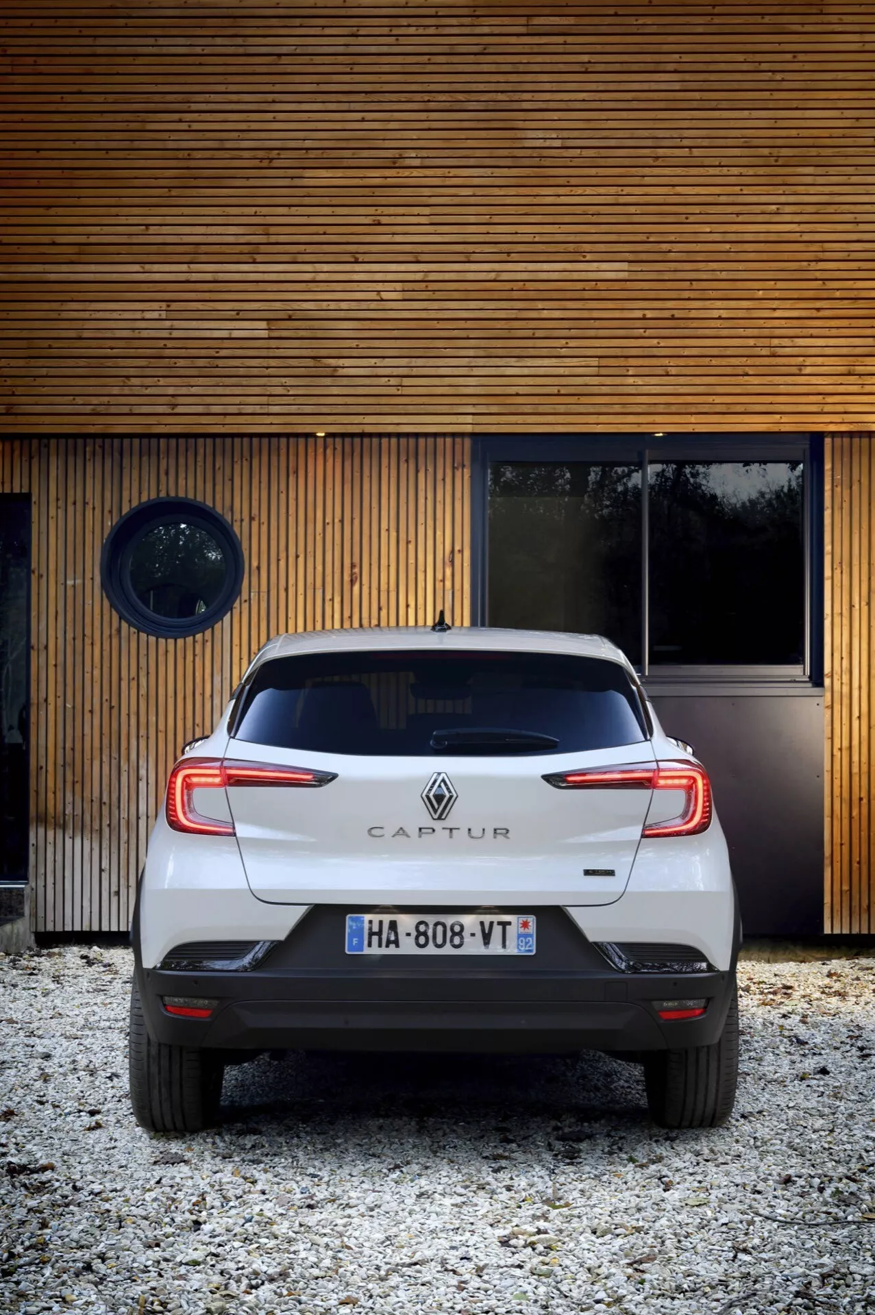 Tylny widok Renault Captur 2024 (lifting II), kompaktowego crossovera, pokazujący tylne światła, zderzak, klapę bagażnika i tablicę rejestracyjną z niskiej perspektywy w górę, z modernistycznym tłem i oświetleniem dziennym