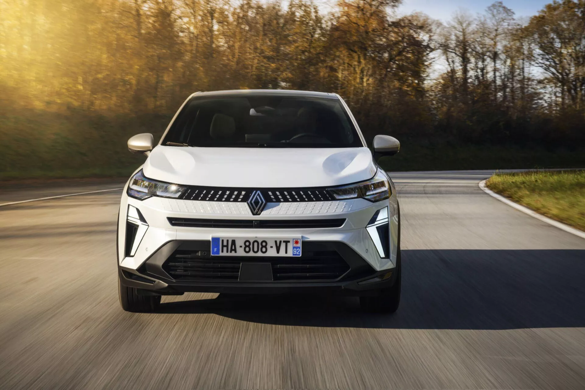 Facelifting Renault Captur 2024 w ruchu z niskiego kąta widzenia frontalnego, podkreślający jego dynamiczny projekt i nowoczesne cechy, w tym LED-owe reflektory, elegancka atrapa z centralnym znaczkiem i aerodynamiczny zderzak przedni, na tle jesiennego krajobrazu w ruchu