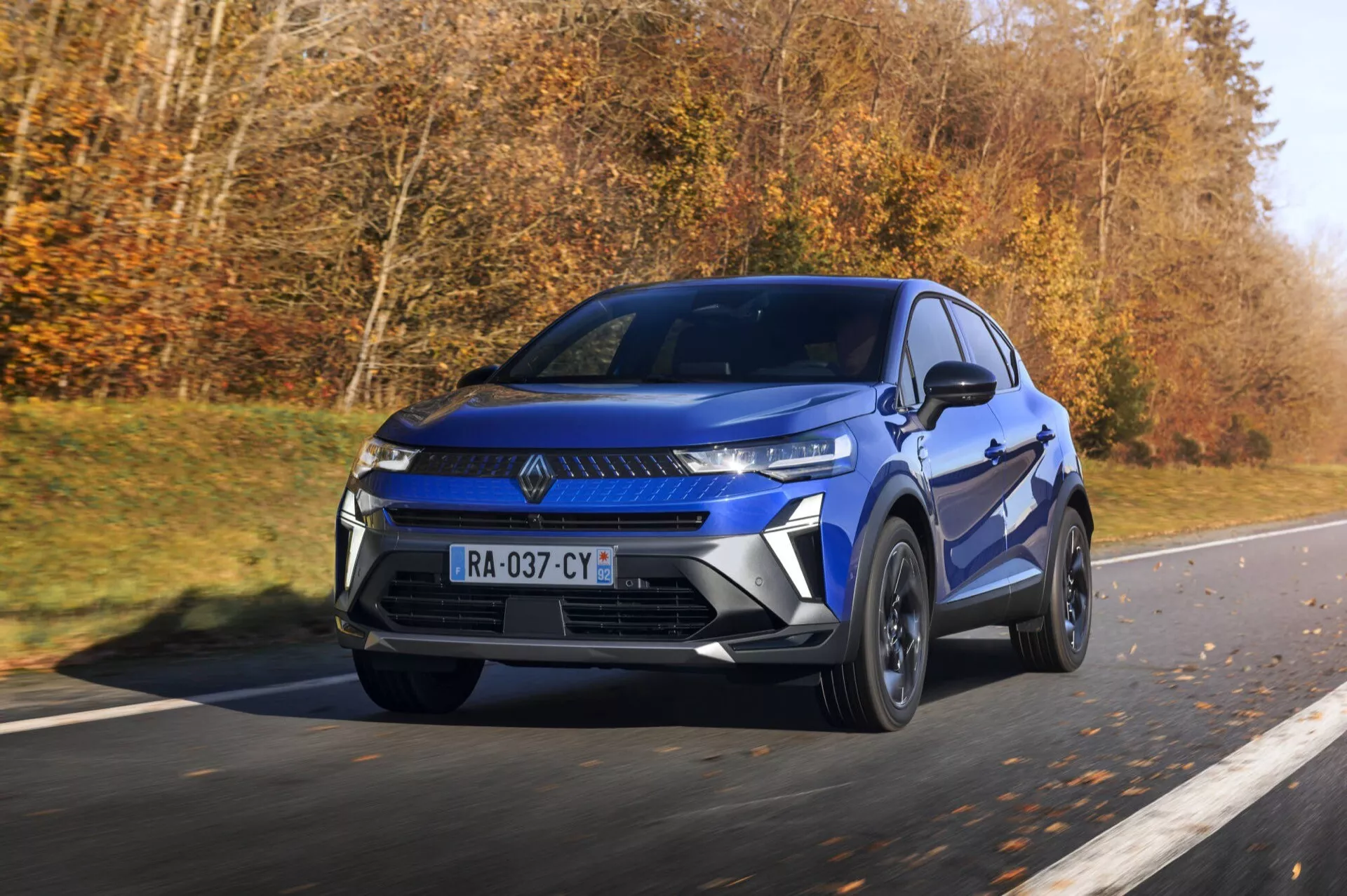 Renault Captur II po liftingu, rocznik 2024, w ruchu, przedstawiające nowoczesne niebieskie SUV z wąskimi kątowymi reflektorami i wyraźną atrapą na autostradzie, podkreślające sportową elegancję, felgi aluminiowe i dynamiczne detale designu w świetle dziennym.