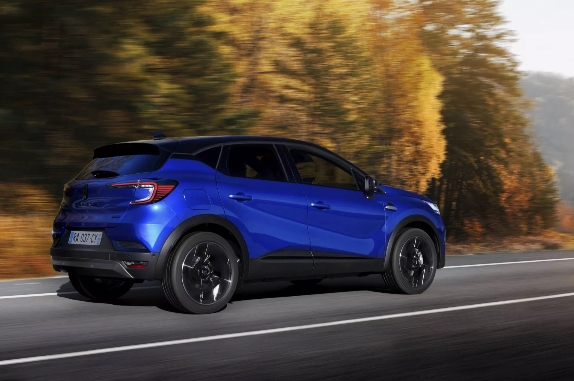 Niebieski sportowy samochód użytkowy Renault Captur 2024 (II-lifting) w dynamicznym ruchu, widok z profilu prezentujący boczne i tylne elementy designu na drodze z jesiennym tłem