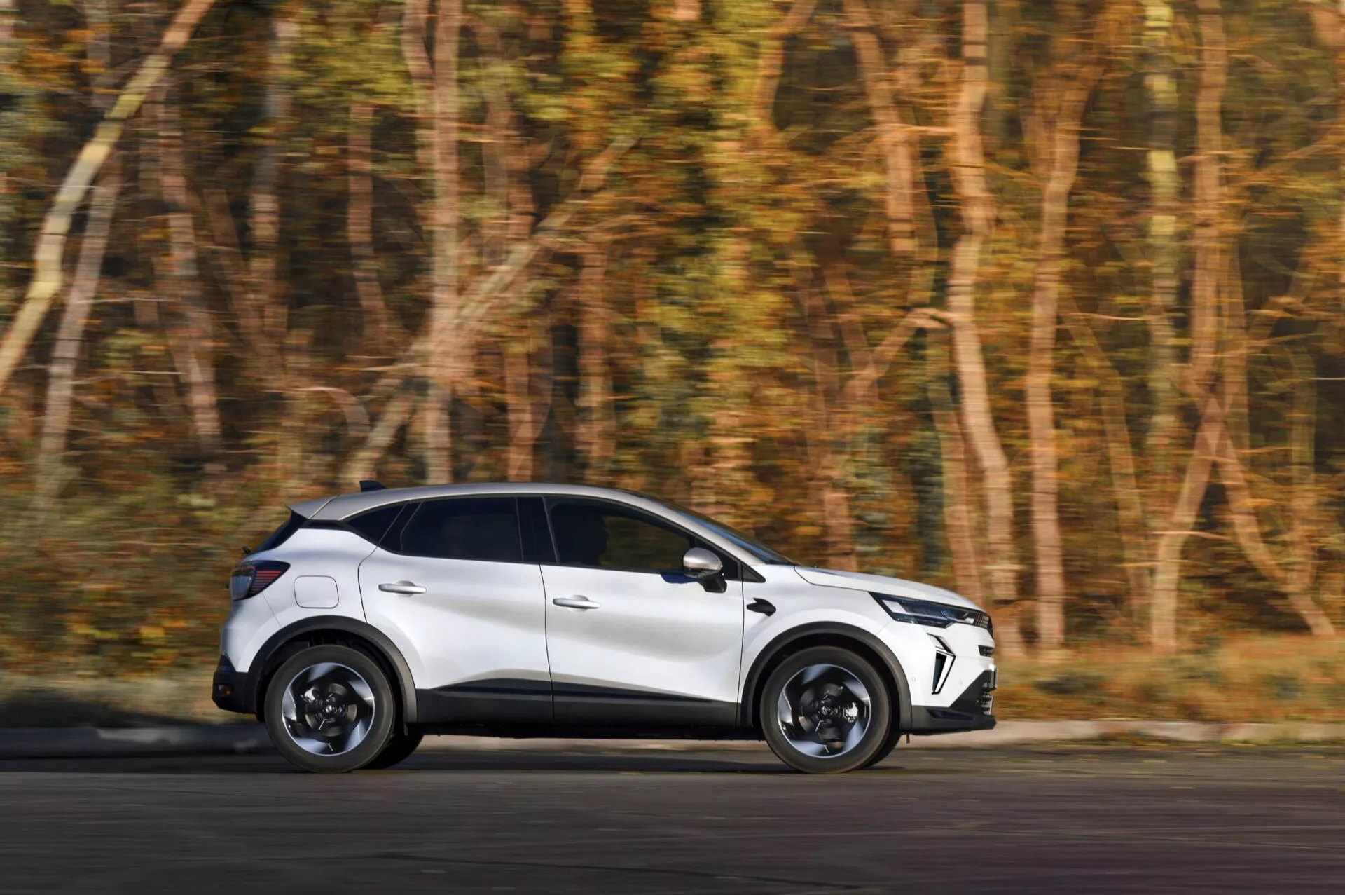 Renault Captur II po liftingu 2024 w ruchu: nowoczesne białe crossover z detalicznym widokiem bocznym i tylnym kątem na zamazanym jesiennym leśnym tle