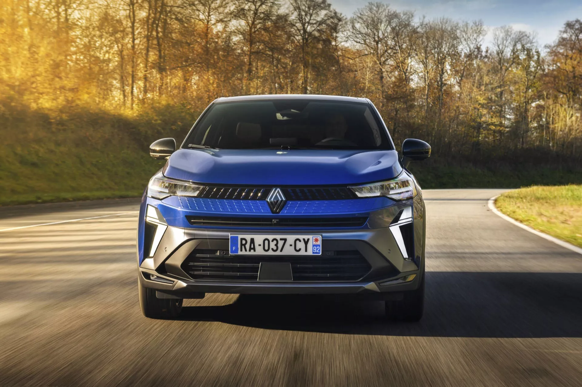 Renault Captur II lifting 2024 w metalicznym niebieskim, pokazujący ostre, kątowe linie, reflektory LED z światłami do jazdy dziennej, dużą kratę przednią i nowoczesny design aerodynamiczny, uchwycony w ruchu na drodze z rozmytym tłem podkreślającym dynamikę i zaawansowane technologie takie jak czujniki parkowania, widziany pod kątem czołowym trzy czwarte