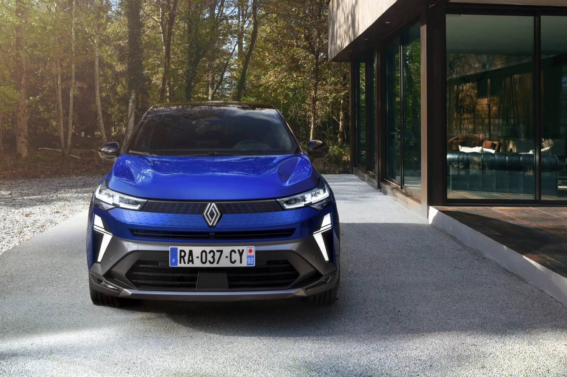 Przedni widok Renault Captur II Facelift 2024 podkreślający eleganckie reflektory, nowoczesną kratę, żywy niebieski lakier i sportowy design zderzaka z zintegrowanymi wlotami powietrza, zaparkowany na podjeździe z tłem nowoczesnego domu