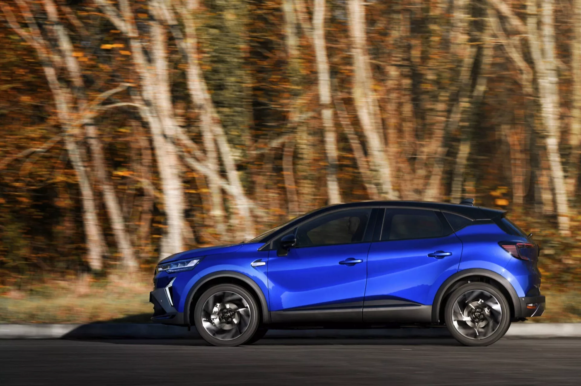 Renault Captur (II-Facelift) 2024 w ruchu, niebieskie SUV z dynamiczną techniką panningu podkreślająca szybkość i detale konstrukcyjne, widok z boku na malowniczej drodze