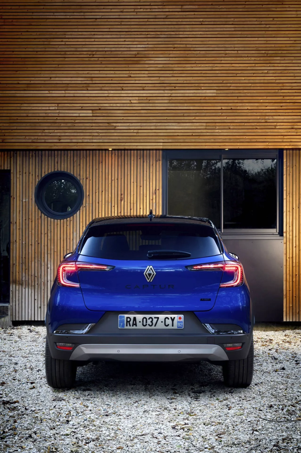 Tylny widok Renault Captur 2024 (II Facelift) na żwirze z nowoczesną drewnianą konstrukcją w tle, prezentujący elementy takie jak tylne światła, zderzak tylny, znaczek i charakterystyczne tylne przednie szyby