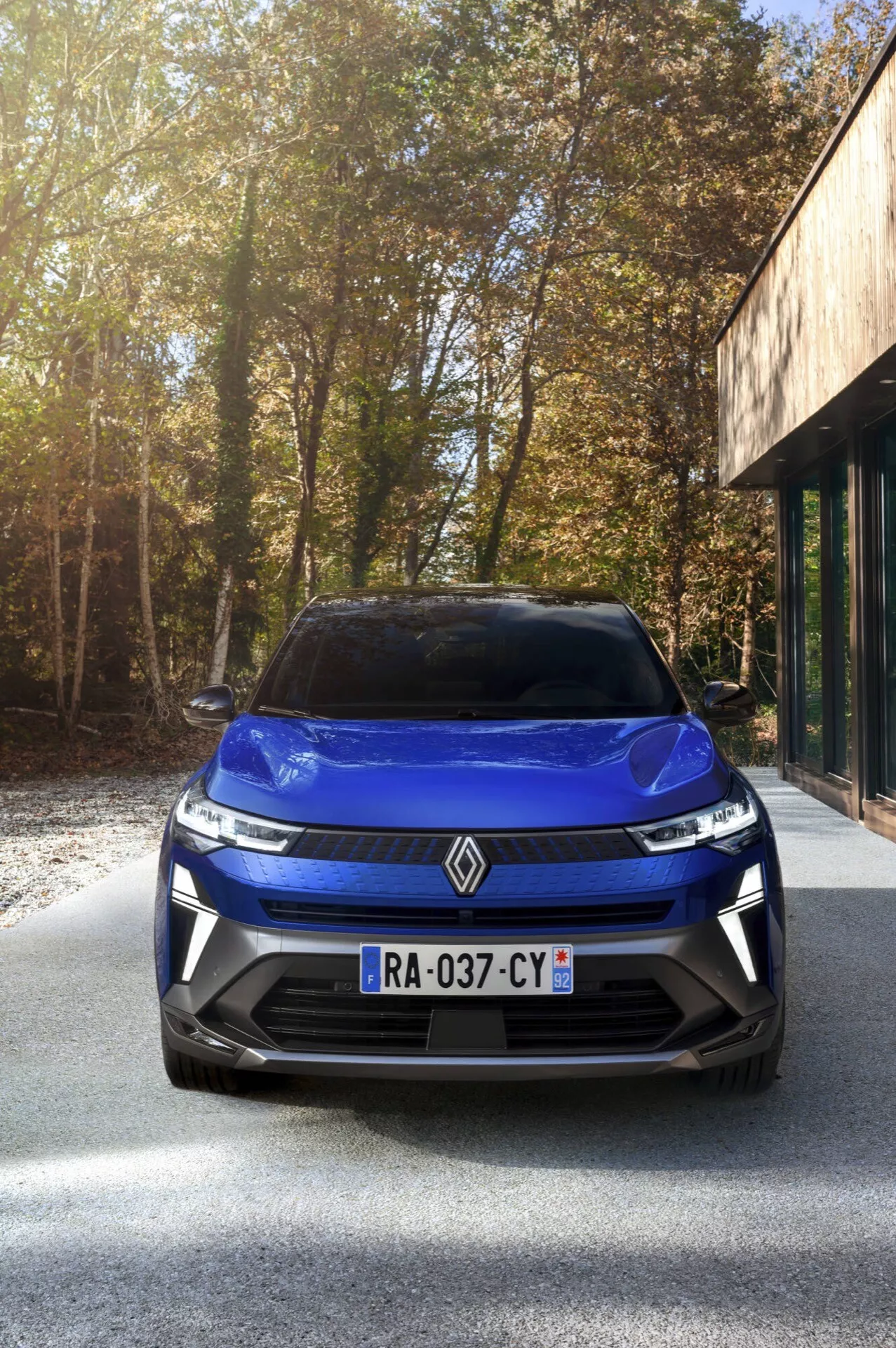 Przód Renault Captur 2024 po liftingu, prezentujący wzór grilla, reflektory LED i emblemat na podjeździe asfaltowym z drzewami i budynkiem z dużymi oknami szklanymi w tle w słonecznych warunkach.