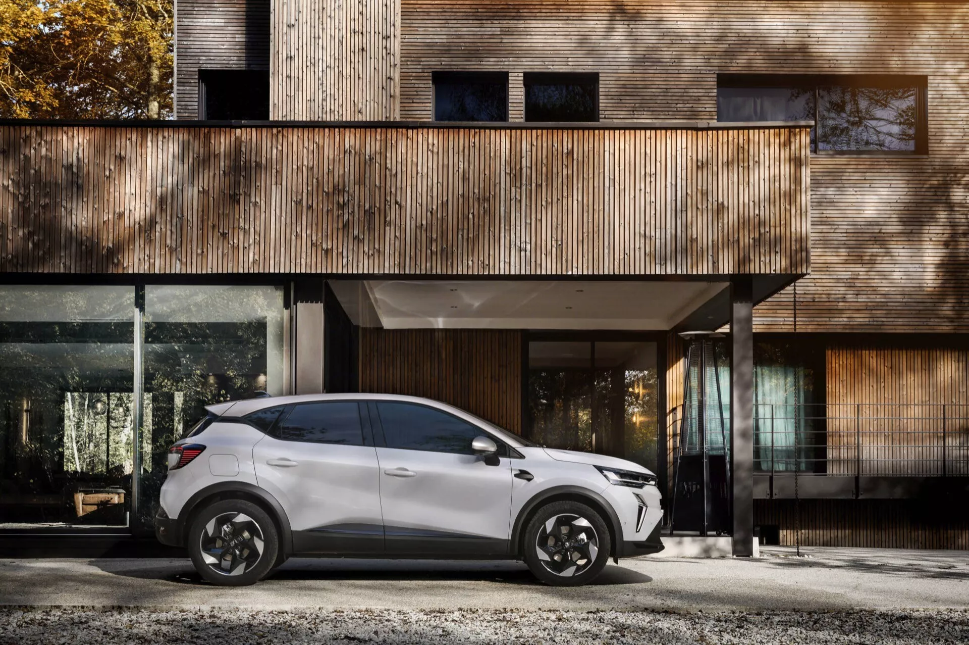 Boczny profil Renault Captur II lifting 2024 zaparkowany przed nowoczesnym domem z drewnianą fasadą i dużymi oknami, stylowe czarne koła ze stopów, elegancki design SUV-a z czterema drzwiami i aerodynamiczne lusterka