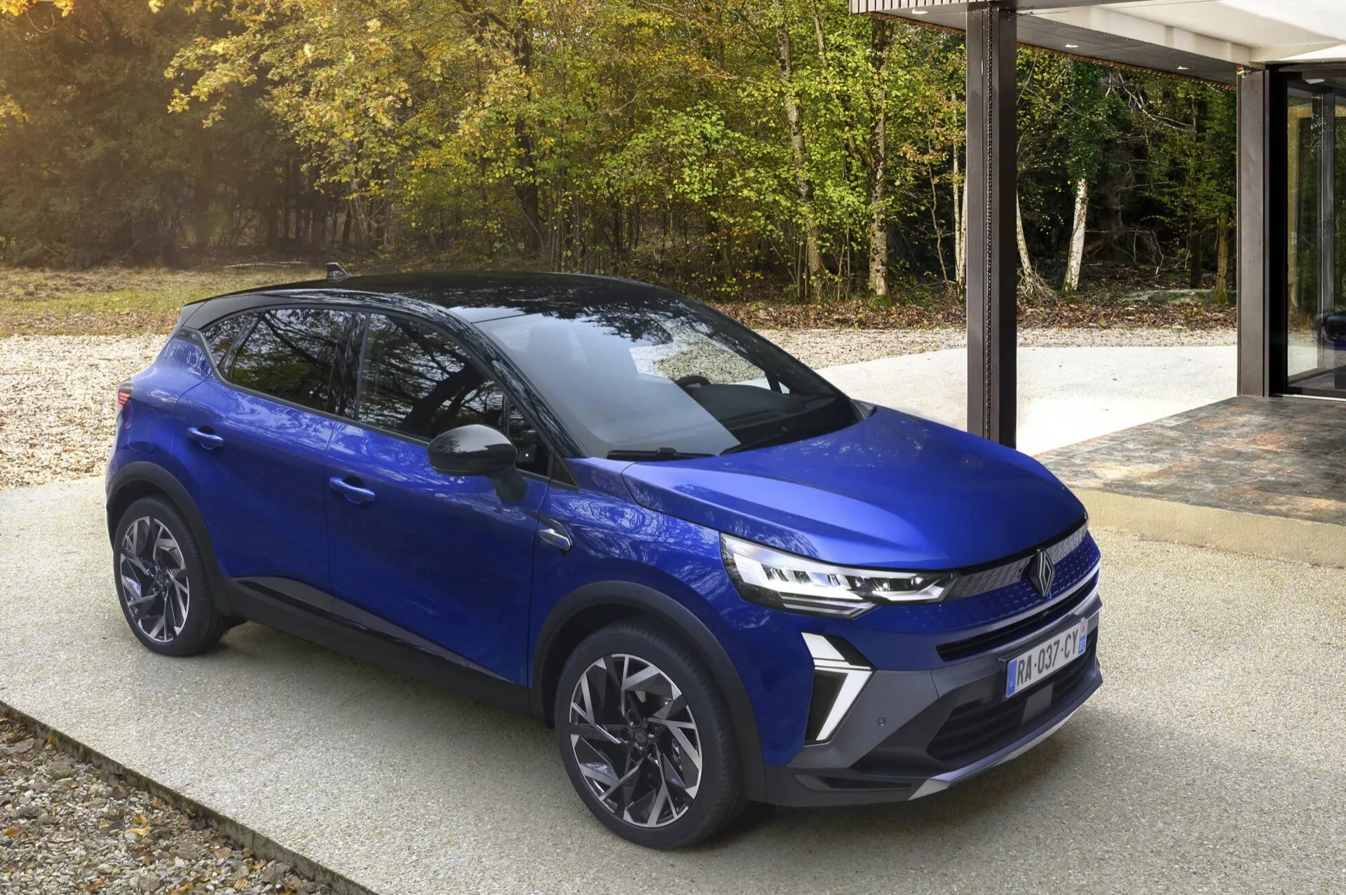 Renault Captur (II-Facelift) 2024 w nowoczesnym niebieskim kolorze, zaparkowany na podjeździe rezydencji z naturalnym tłem drzew i nowoczesnej architektury, prezentujący przedni grill, reflektory LED, felgi aluminiowe i profil boczny w świetle dziennym.