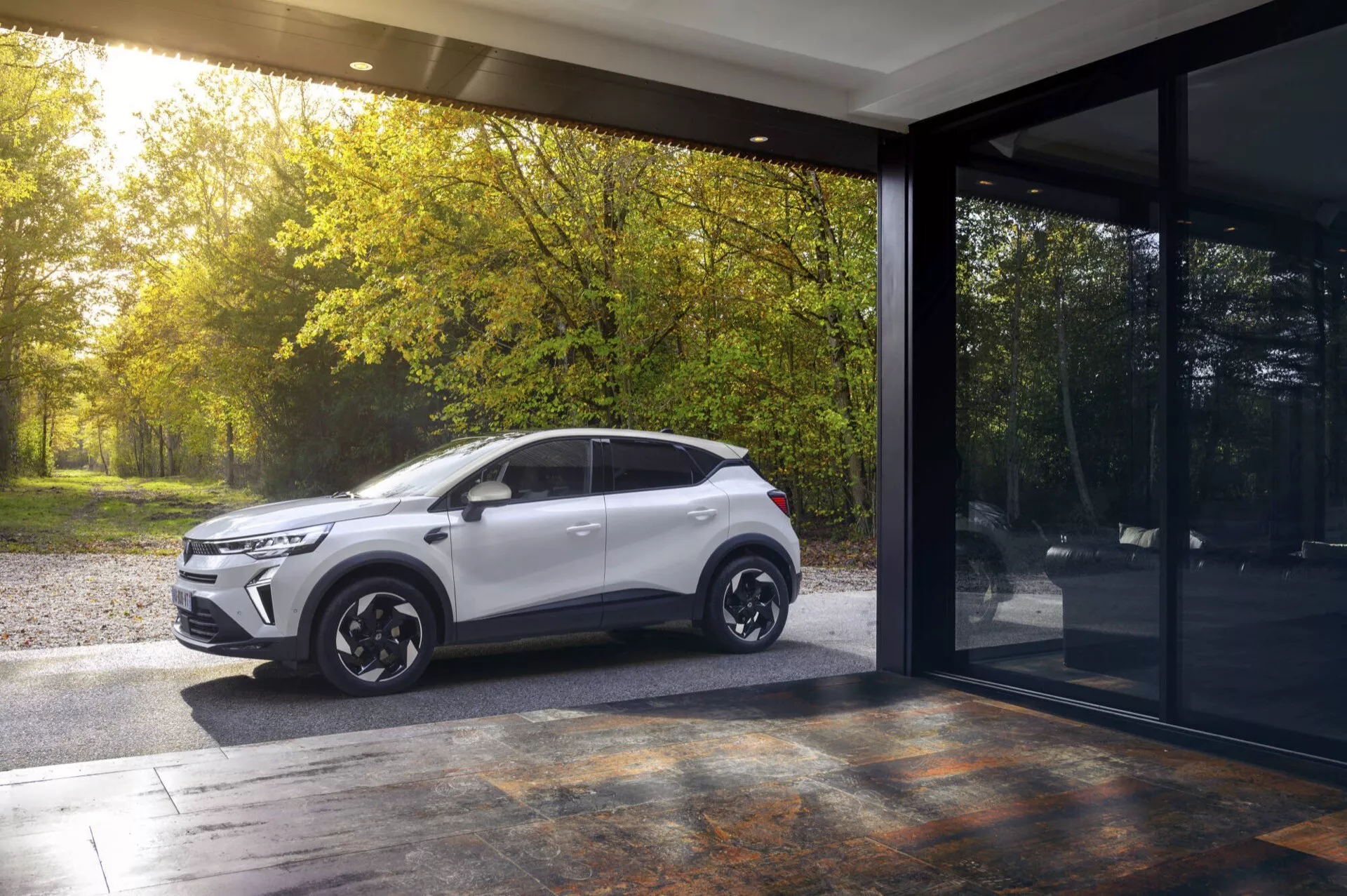Facelifting Renault Captur II z 2024 roku w kolorze białym, zaparkowany na zewnątrz obok budynku z dużymi oknami szklanymi, prezentujący nowoczesny design SUV-a, profil boczny z sugestią przedniej kratki, w świetle słonecznym wczesnej jesieni