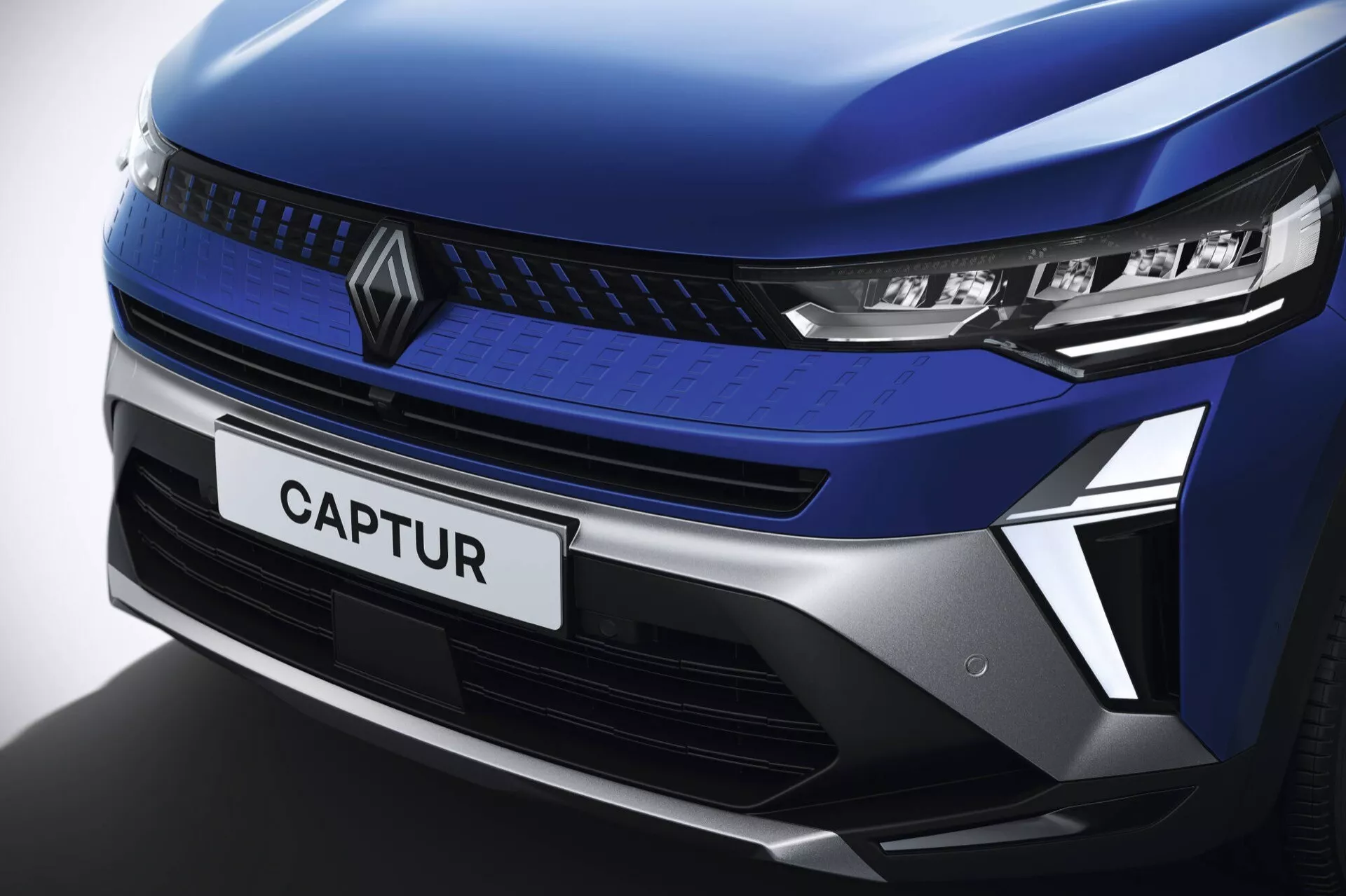 Zbliżenie na liftingowany Renault Captur 2024 z emblematem, elegancką kratką z poziomymi liniami, wąskimi reflektorami LED, nazwą modelu na grillu, metalicznie niebieską maską i zderzakiem z srebrnymi akcentami, skupiając się na przedniej części pojazdu i aerodynamicznej estetyce.