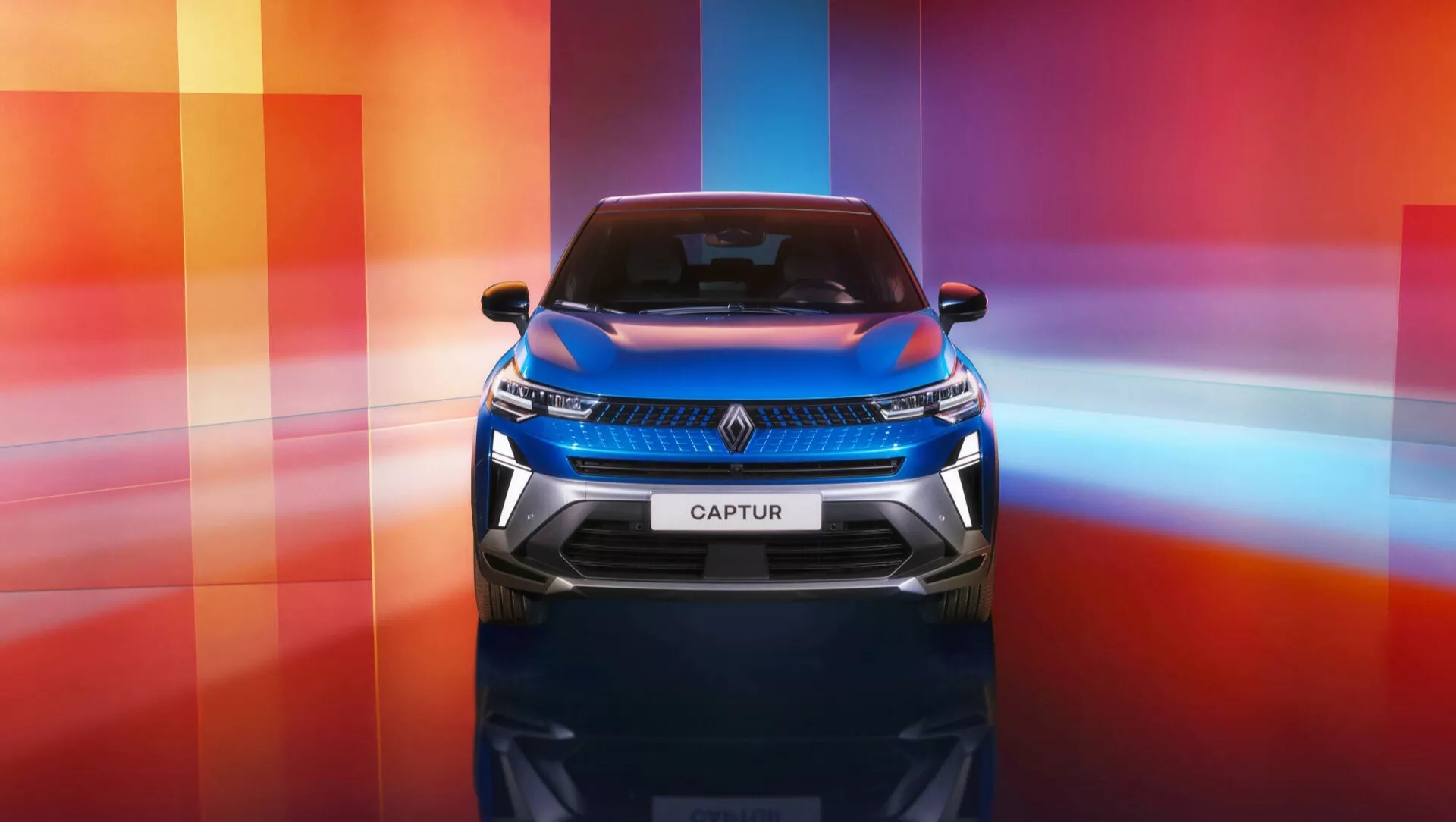 Renault Captur II Facelift 2024 prezentowany z frontowej perspektywy podkreślającej tabliczkę 'CAPTUR' na przednim grillu, reflektory LED z charakterystycznym podpisem świateł do jazdy dziennej, nowoczesny design grilla, potencjalne punkty integracji systemów ADAS na zderzaku i grillu, błyszczące wykończenie, na dynamicznym wielokolorowym tle.