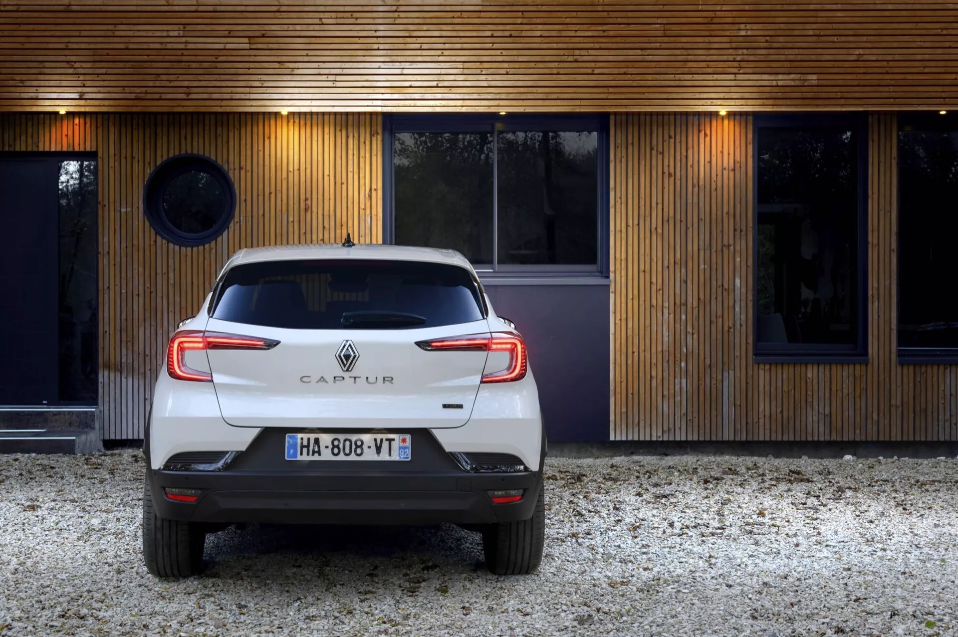 Tylny widok białego Renault Captur 2024 (II-facelift) zaparkowanego przed nowoczesnym budynkiem z drewnianą fasadą, prezentujący tylne światła LED w kształcie litery C, emblemat i czujniki parkowania.