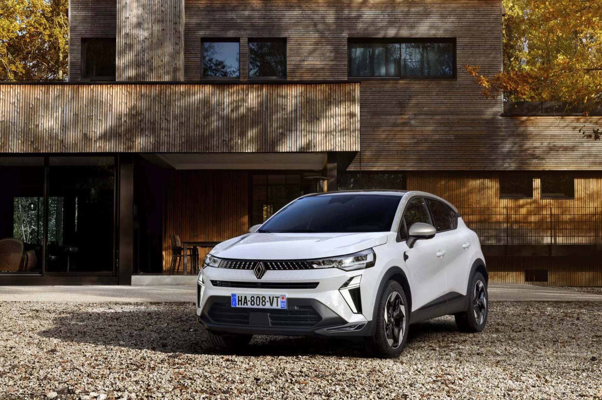Zmodernizowany model Renault Captur 2024 zaparkowany pod kątem, prezentujący nowoczesny design SUV, z wyraźną przednią kratką z poziomymi liniami, smukłymi reflektorami z diodami LED, aerodynamicznie ukształtowaną maską i sportowym, opadającym dachem na tle luksusowej zabudowy mieszkalnej