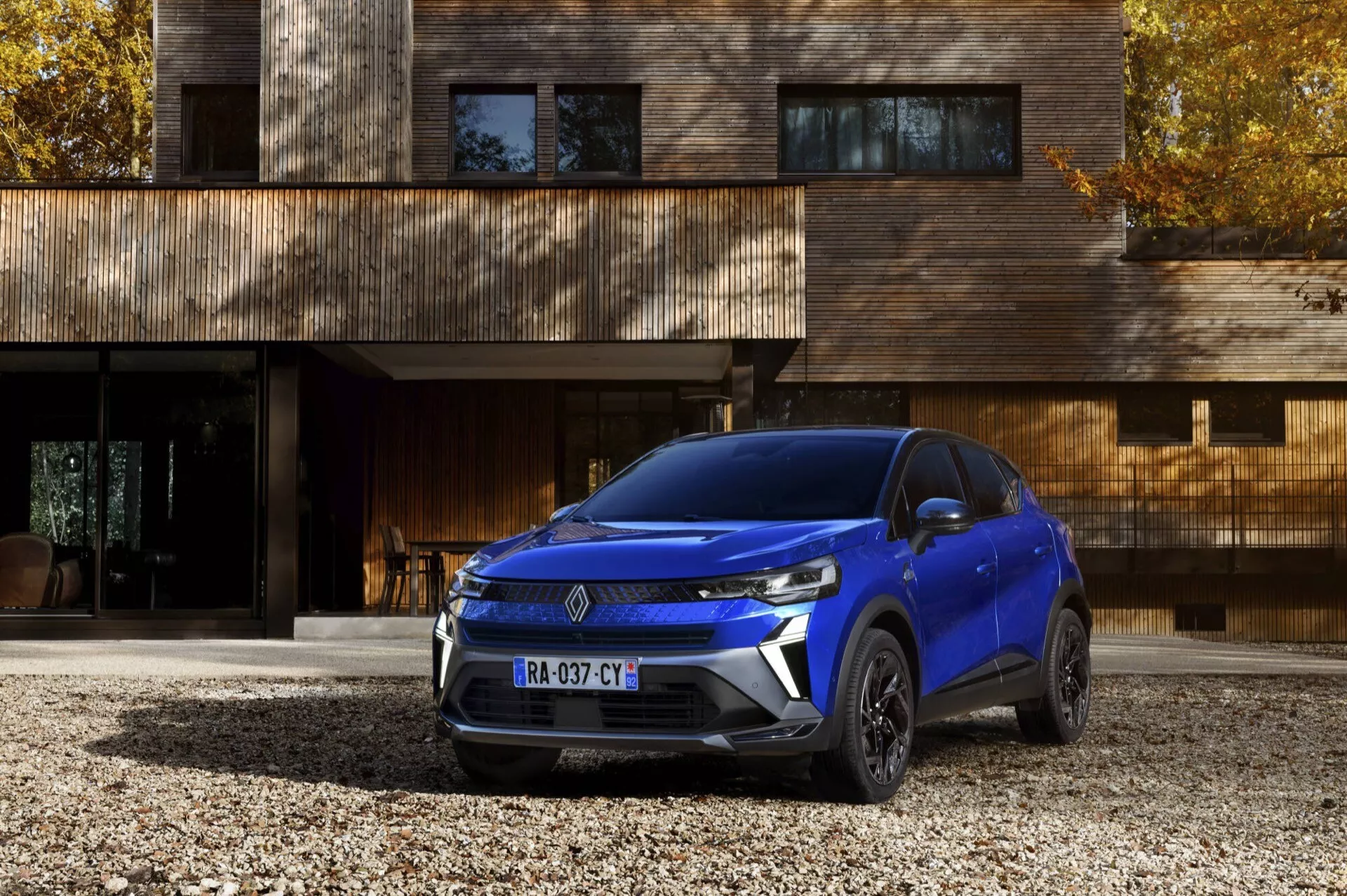 Niebieski SUV Renault Captur (II Facelift) 2024 zaparkowany na żwirowym podjeździe przed nowoczesnym drewnianym domem, prezentujący swoje eleganckie reflektory LED, kątową przednią atrapę i złożone felgi aluminiowe z niskiej kwartalnej perspektywy przedniej
