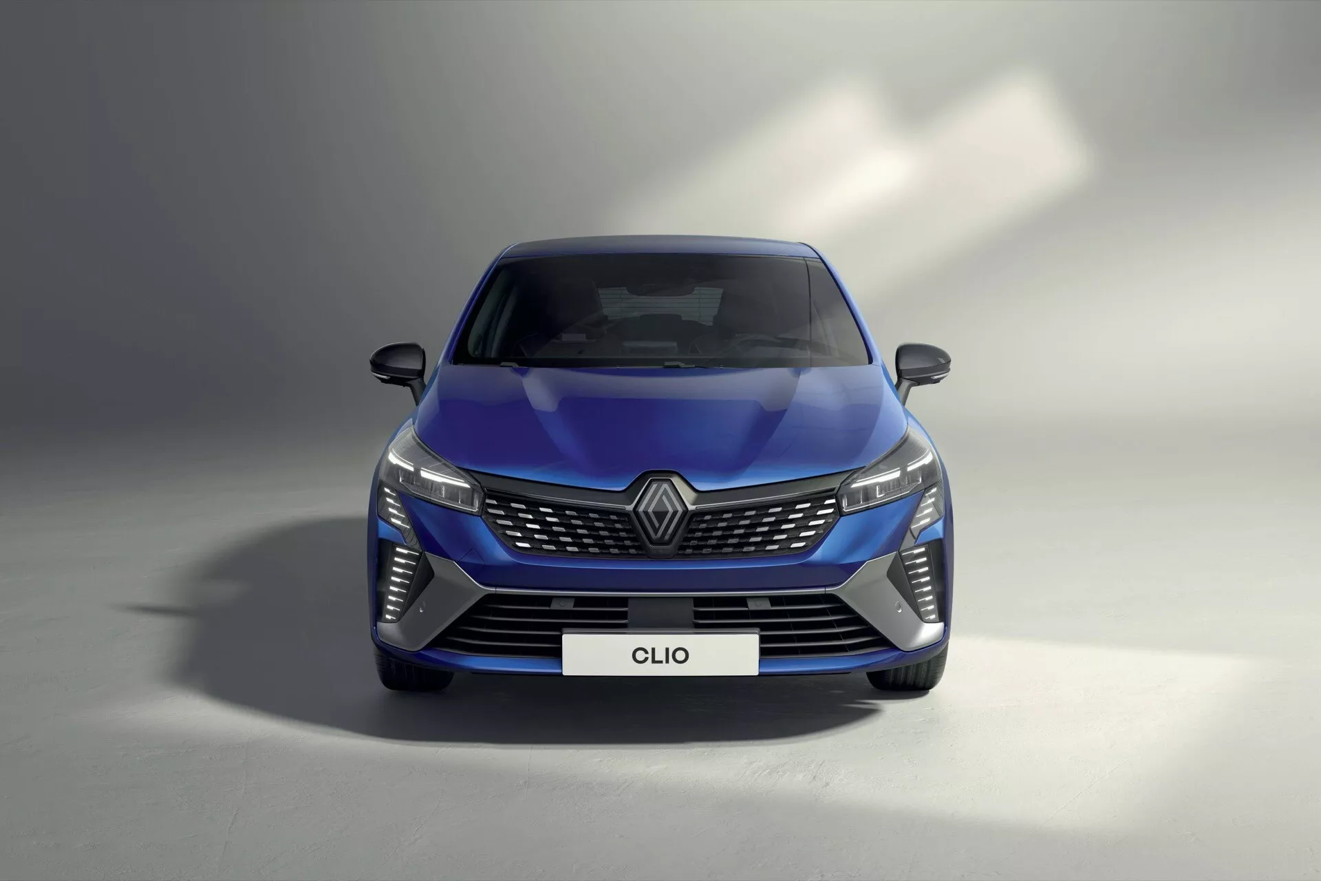 Renault Clio V Facelift 2023 w kolorze niebieskim, pokazujący LEDowe reflektory, skomplikowany wzór grilla, aerodynamiczny przedni zderzak i zaawansowane lusterka bezpieczeństwa z niskiego kąta przedniego, podkreślający technologię oświetlenia samochodowego, elementy brandingowe i aerodynamiczny styl.