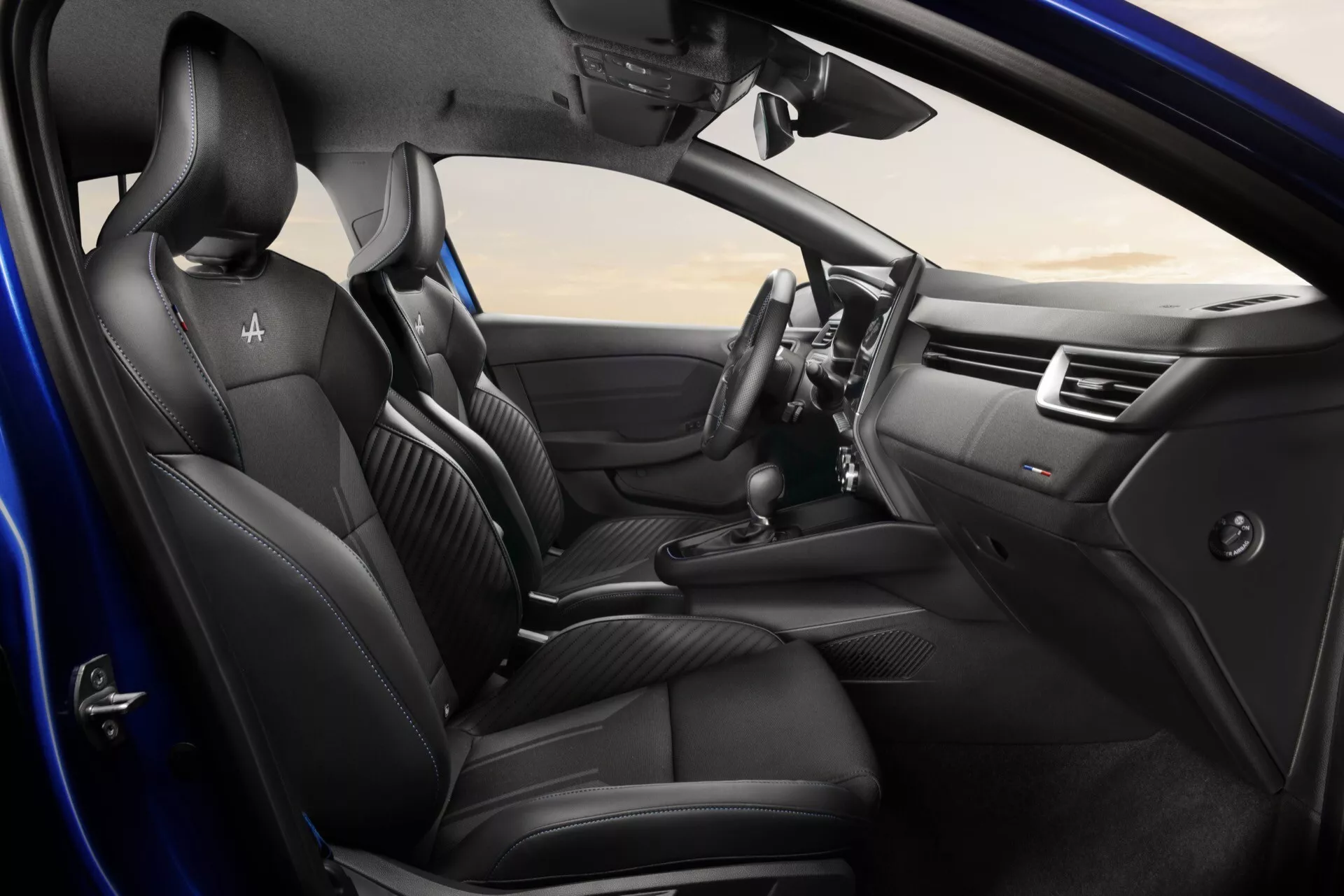 Wnętrze Renault Clio 2023 (V-Facelifting) z czarnymi skórzanymi przednimi siedzeniami, wielofunkcyjną kierownicą i centralnym systemem infotainment, widziane z lekko podniesionego kąta z otwartymi drzwiami po stronie kierowcy i szczegółami deski rozdzielczej
