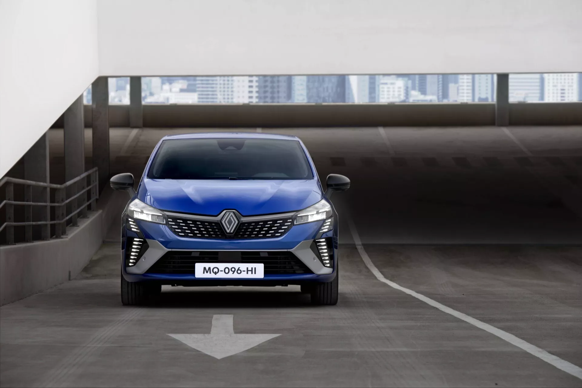 Niebieski Renault Clio V-facelift 2023, widok z przodu na piętrowym parkingu z kierunkową strzałką, prezentujący reflektory LED, wyrazistą grillę z dużym logo i sportowy design zderzaka przedniego