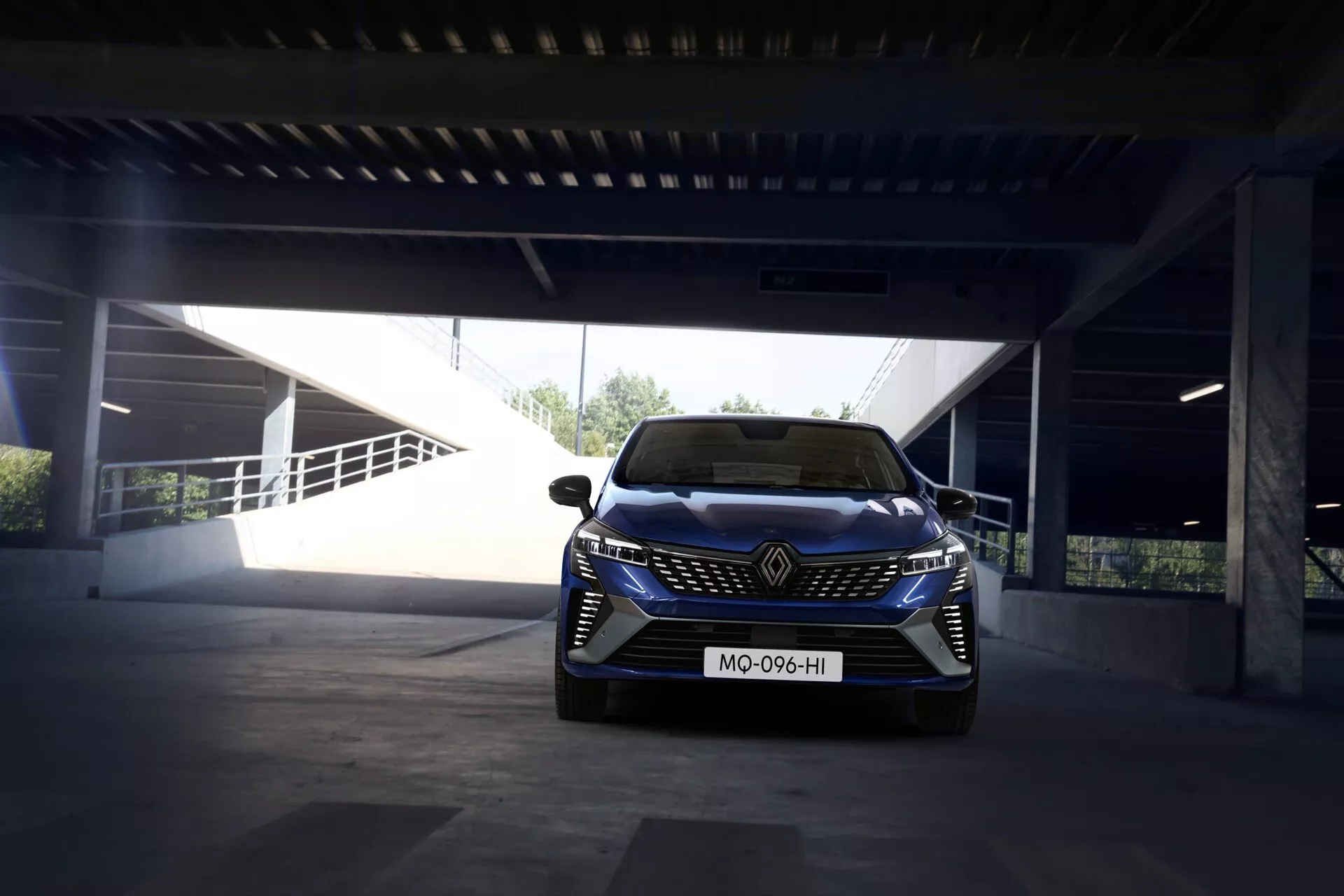 Renault Clio V-Facelift 2023 w dzień na parkingu; widok z przodu pokazujący emblemę marki, reflektory LED, sportowy zderzak z wlotem powietrza i czujniki wspomagające kierowcę.