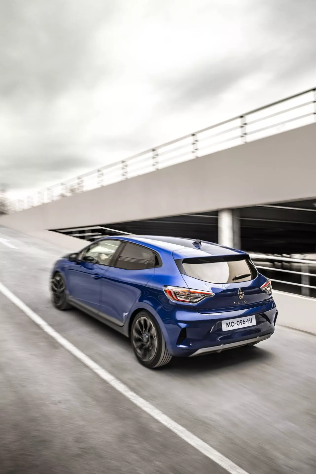 Dynamiczny widok z tyłu ukośny Renault Clio V-Facelift 2023 w ruchu na drodze, pokazujący sportowy design, tylne światła LED, spojler montowany na dachu i koła wieloramienne
