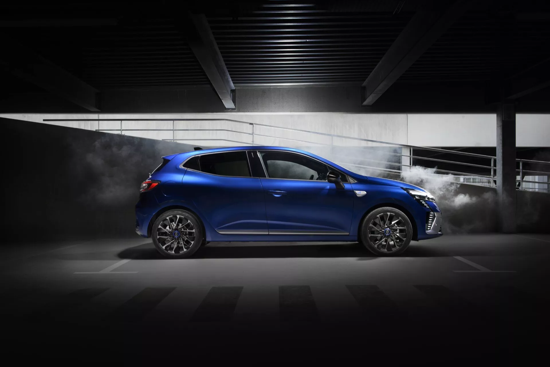 Renault Clio V-Facelift 2023 w lśniącym niebieskim kolorze z dynamicznym profilem bocznym, zdjęcie z niskiego kąta podkreślające sportową postawę, aerodynamiczne linie projektowe, felgi ze stopu o wielu szprychach i efekty sugerujące szybkość, w dramatycznie oświetlonym wnętrzu