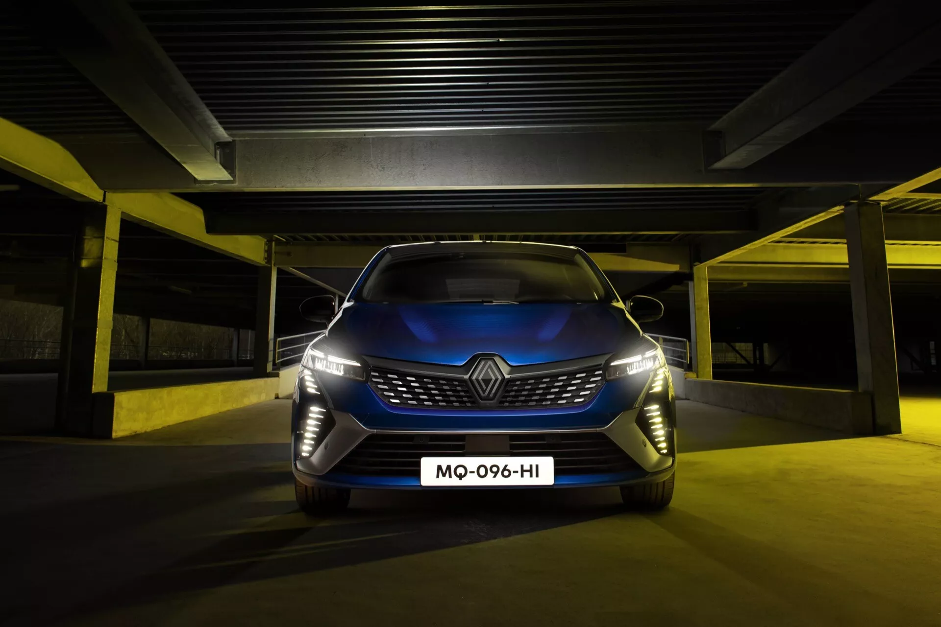Renault Clio V-Facelift 2023 prezentuje swoją odważną atrapę i zaawansowane reflektory LED z niskiego kąta przedniego, podkreślając zaawansowane cechy projektowe i technologiczne, w scenerii słabo oświetlonego parkingu.