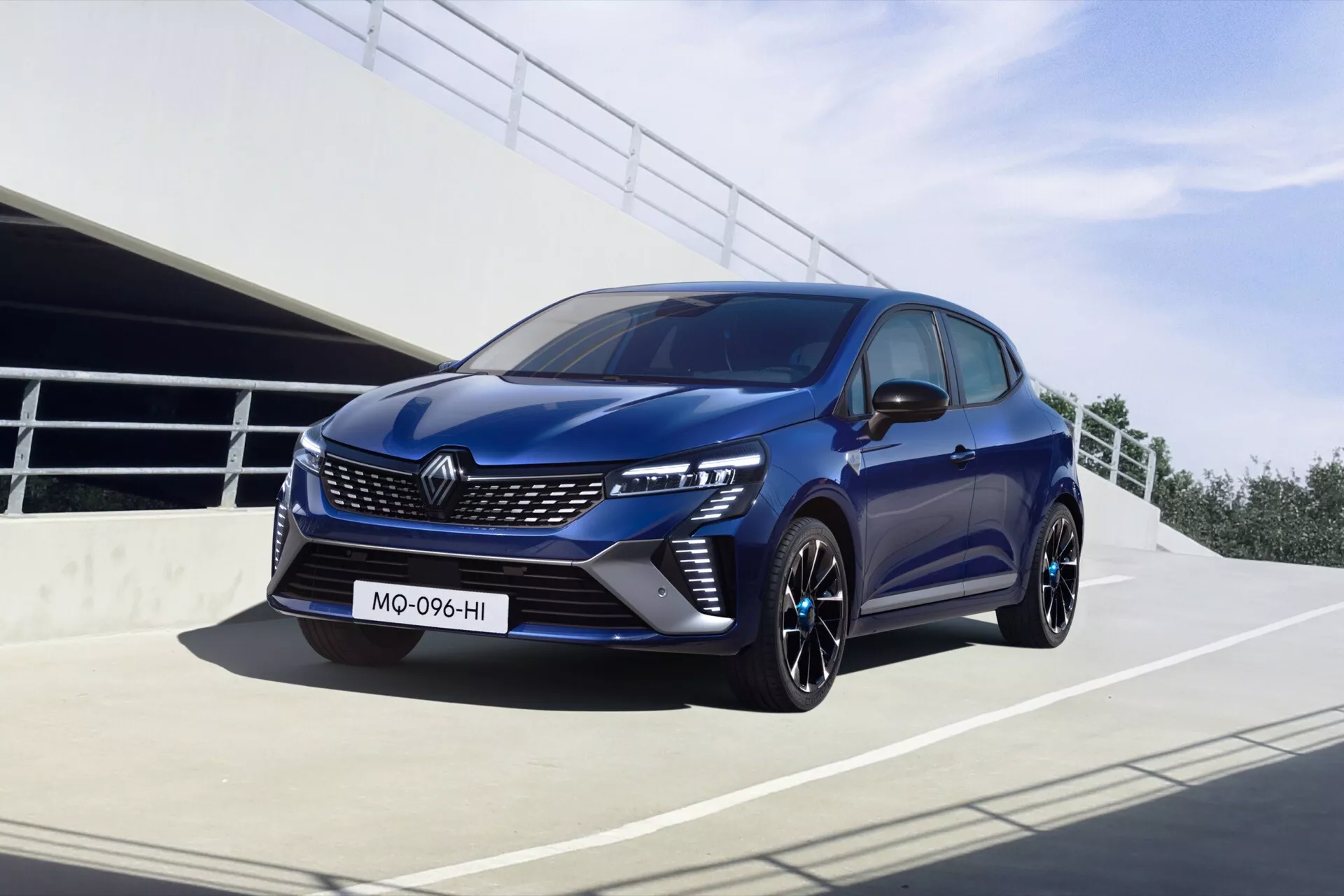 Nowoczesny hatchback Renault Clio V Facelift 2023 w dynamicznej pozie przedstawiający przód pojazdu, reflektory LED, sportowy zderzak z wlotami powietrza, felgi aluminiowe i głębokie niebieskie malowanie nadwozia, sfotografowany w miejskim otoczeniu