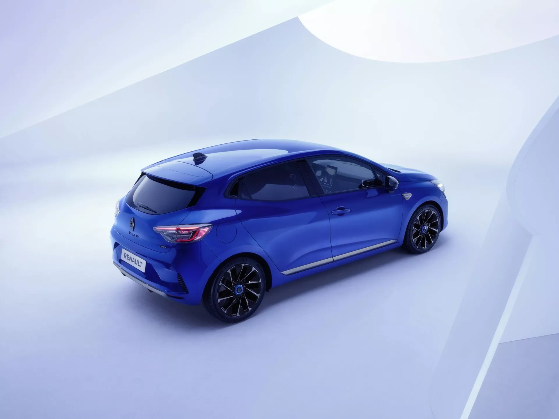 Widok z wysokiego kąta z tyłu trzech czwartych na niebieskie hatchback Renault Clio V-facelift 2023, podkreślający jego tylne światła LED, czujniki parkowania z tyłu, sportowe koło ze stopu i zintegrowaną kamerę cofania na tle łagodnego gradientu