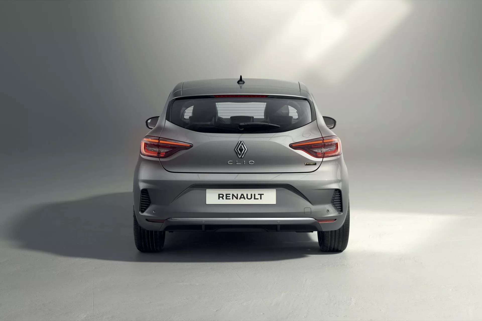 Tylny widok hatchbacka Renault Clio 2023 prezentujący C-kształtne diody LED w tylnych światłach, centralne logo Renault oraz oznaczenie modelu Clio na klapie bagażnika, tylną szybę z funkcją rozmrażania, zintegrowane odblaski i czujniki parkowania w zderzaku tylnym, ukryty design systemu wydechowego, częściowy widok na koła ze stopów lekkich, metaliczny szary lakier z gradientowym cieniowaniem, oraz antenę w kształcie płetwy rekina, pokazujący postęp w projektowaniu samochodów, technologii oświetlenia LED i funkcjach wspomagania kierowcy.