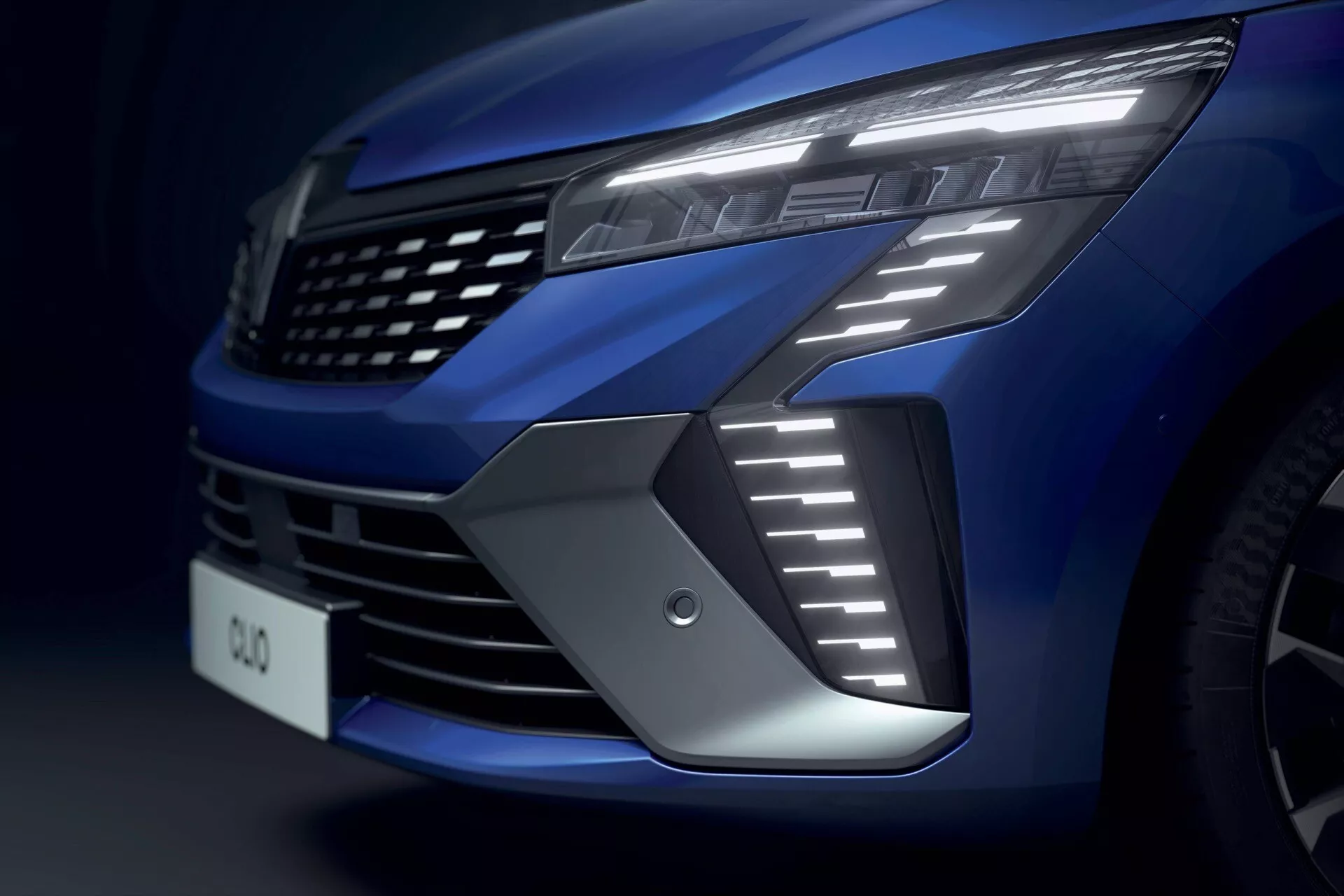 Zbliżenie na przednią część Renault Clio v-facelift 2023, wyróżniające lewą stronę grilla, nowoczesne światło LED z ostrymi światłami dzienymi, wlot powietrza i metaliczne, głębokie niebieskie wykończenie