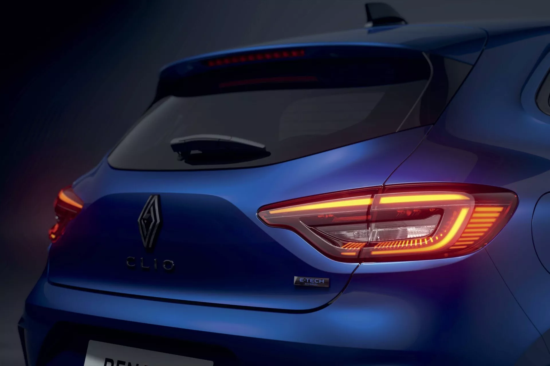 Zbliżenie na Renault Clio V-facelift 2023 pokazujące podświetlony tylny światło w kształcie litery C, szczegół znaczka i elegancki tylny design w metalicznie niebieskim kolorze pod oświetleniem studyjnym