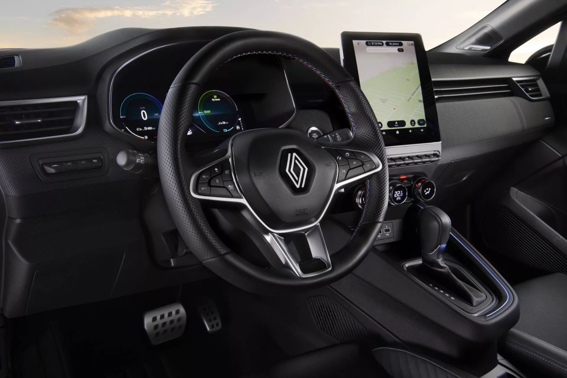 Wnętrze Renault Clio V-Facelift 2023 z widokiem na kierownicę z logo i przyciskami wielofunkcyjnymi, cyfrowy zestaw wskaźników, pionowy system infotainment z dotykowym ekranem i nawigacją, panel sterowania klimatyzacją, nowoczesną dźwignię zmiany biegów, oraz elegancki, minimalistyczny czarny design z perspektywy pasażera