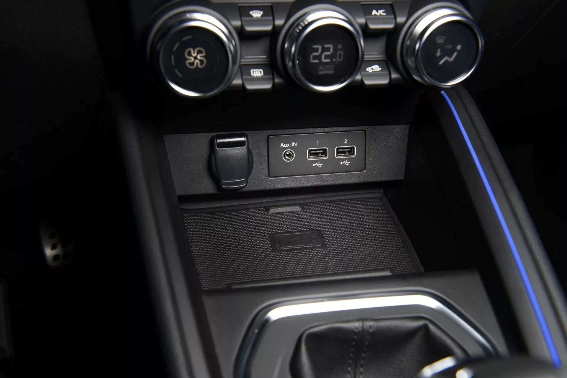 Zbliżenie na środkową konsolę Renault Clio V-Facelift 2023 z pokrętłami kontroli klimatyzacji, portami USB i AUX, gniazdem zasilania 12V, zatopioną przegródką na drobiazgi i obszarem dźwigni zmiany biegów z niebieskim oświetleniem ambientowym