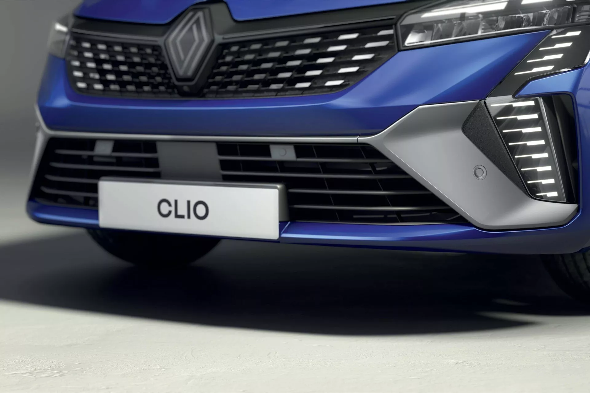 Zbliżenie, widok z niskiego kąta na przednią część Renault Clio V-facelift 2023, skupiający się na grillu, wlotach powietrza i designie zderzaka, prezentujący błyszczące metaliczne niebieskie wykończenie i zaawansowane oświetlenie