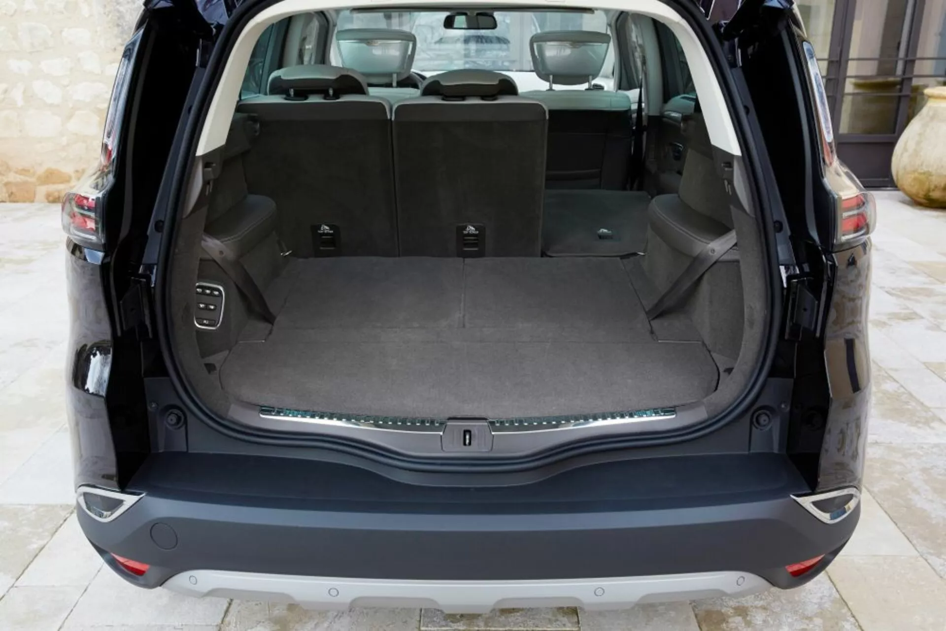 Tylny widok wnętrza Renault Espace 2015 z czystym obszarem ładunkowym i otwartą klapą, przedstawiający płaską powierzchnię ładunkową, tylną kanapę dzieloną w stosunku 60/40, gniazdo zasilające, haki ładunkowe, przycisk sterowania elektronicznie otwieraną klapą bagażnika oraz chromowaną listwą ozdobną, przy naturalnym oświetleniu.