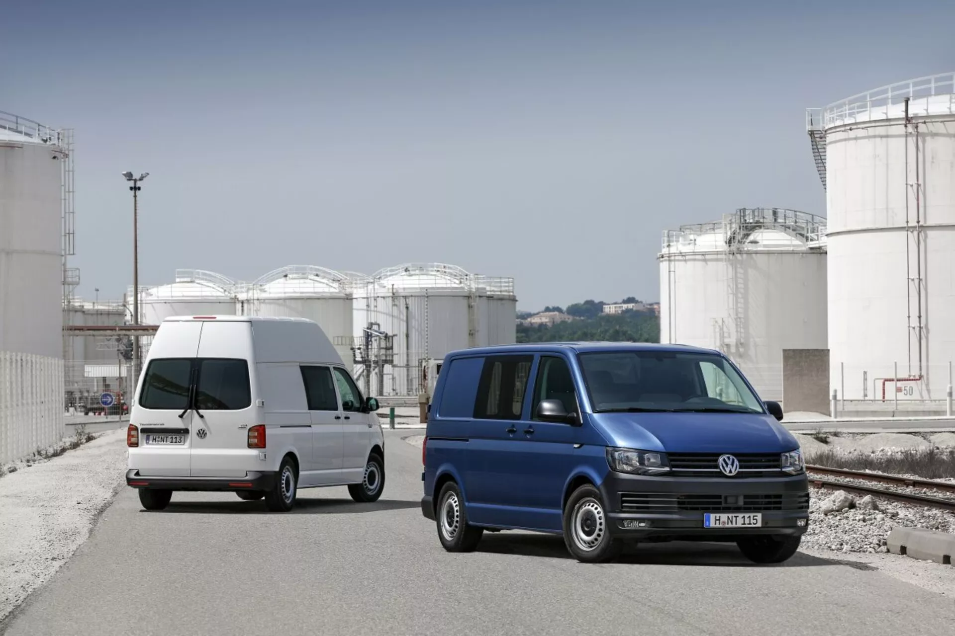 Dwa vany Volkswagen Transporter zaparkowane na terenie przemysłowym z dużymi zbiornikami magazynowymi w tle, prezentujące ich użytkowy design komercyjny, ślizgowe drzwi boczne i standardowe koła ze stopu.