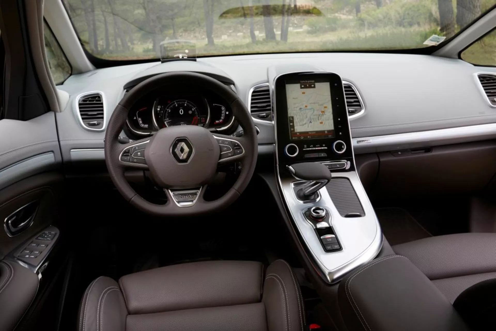 Widok wnętrza Renault Espace 2015 z perspektywy kierowcy, przedstawiający luksusowe wykończenie z kierownicą trójramienną z przyciskami sterowania, cyfrowym zegarem, pionowym ekranem dotykowym konsoli środkowej do systemu infotainment, automatyczną dźwignią zmiany biegów, siedzenia tapicerowane skórą z elektroniczną regulacją i nawiewy klimatyzacji na desce rozdzielczej