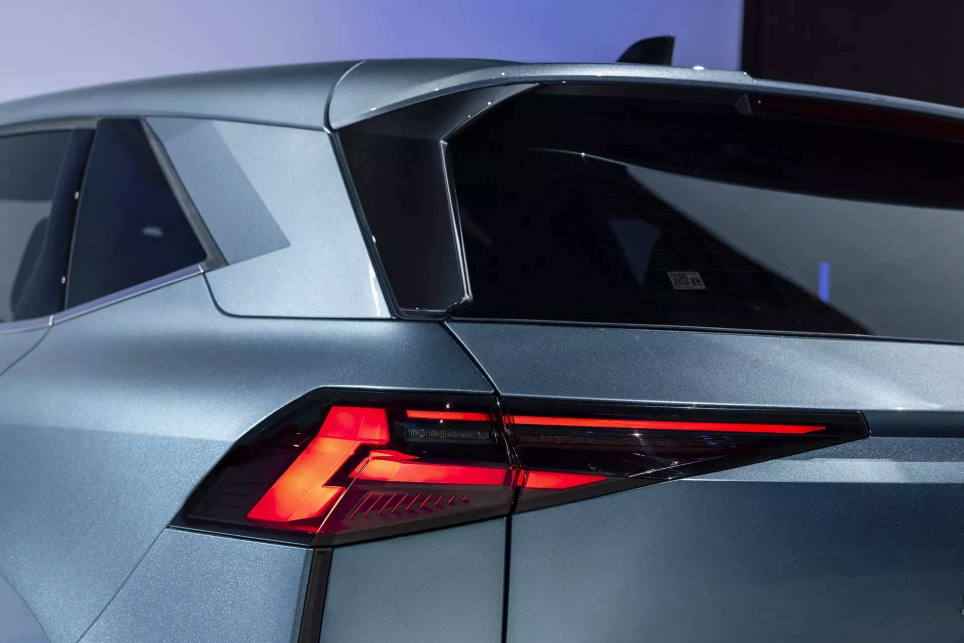 Zbliżenie nowoczesnego Renault Symbioz 2024 (1. generacji) z tylnego, trzyczwartowego kąta. Obraz wyraźnie prezentuje ostry, kanciasty design świateł LED, nachyloną tylną szybę i charakterystyczną linię dachu z małym spojlerem. Nadwozie ukazuje futurystyczne kształty geometryczne w metalicznym, srebrnym/szarym wykończeniu. Na lewej stronie bagażnika widoczny jest znaczek lub emblemat, sugerujący model lub markę samochodu. Inne szczegóły obejmują część przypominającą wlewu paliwa na lewym tylnym panelu.
